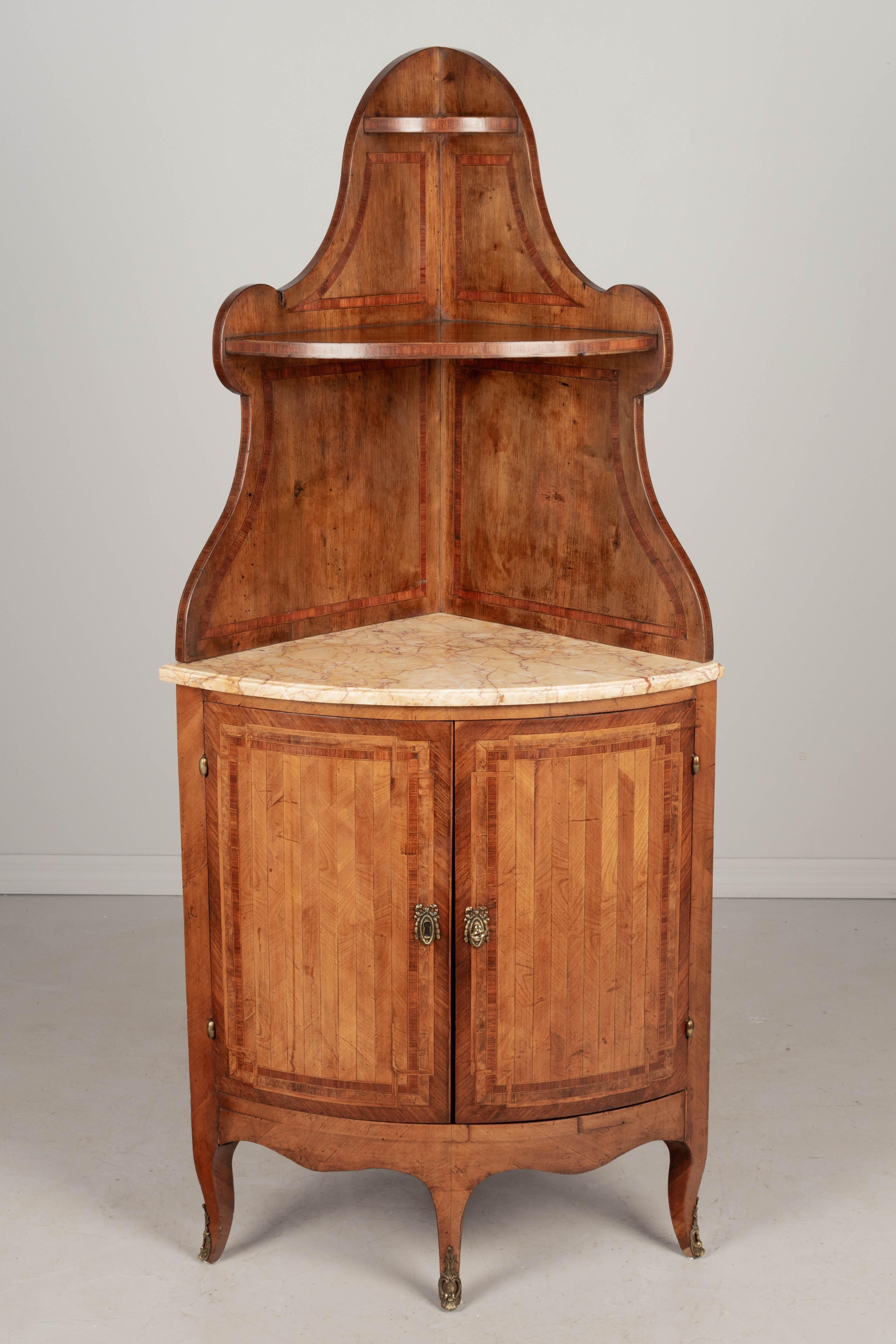A 19th century French Louis XVI style marquetry corner cabinet with inlaid veneers of mahogany, walnut and cherry. In two parts, the marble top demilune cabinet opens to one interior shelf and has a working lock and one key. The tiered shelf rests