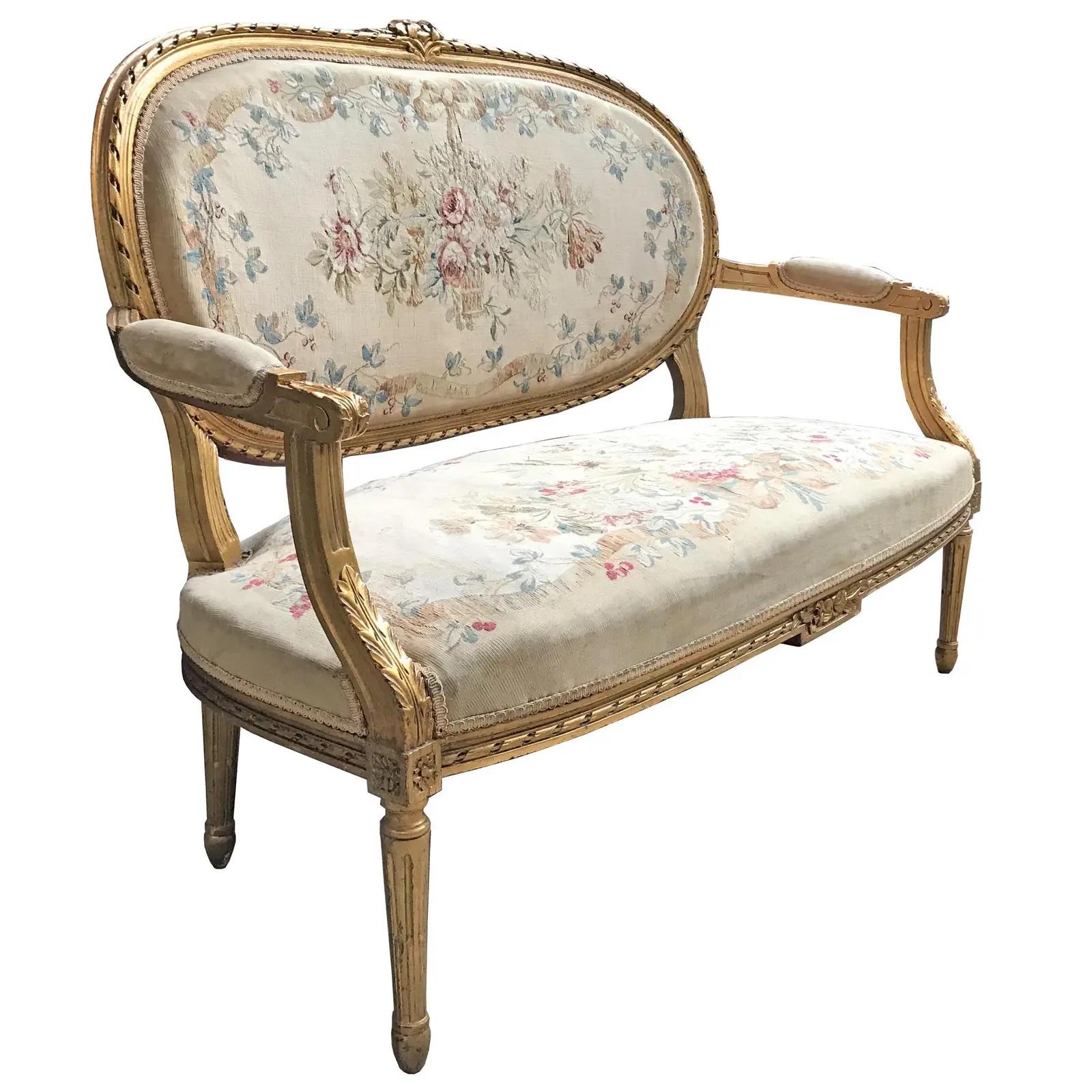 A 19th century French Louis XVI style giltwood settee with a floral cartouche at the top of the back, and circled in a carved ribbon motif, with fluted and tapered legs. The back and seat are upholstered in a floral Aubusson with large-scale