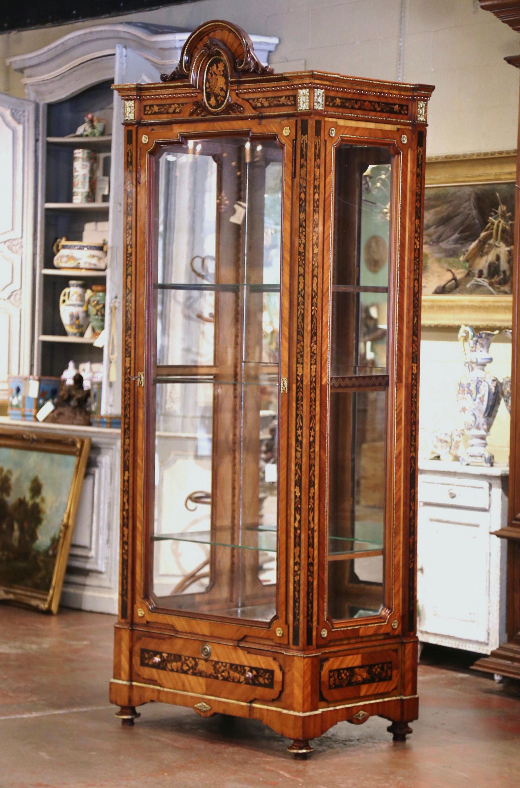 This elegant, antique fruitwood display cabinet was crafted in France, circa 1880. The tall vitrine stands on bun feet decorated with bronze mounts; the top features a crest, which is centered within a floral inlaid, ebonized panel. The same inlay