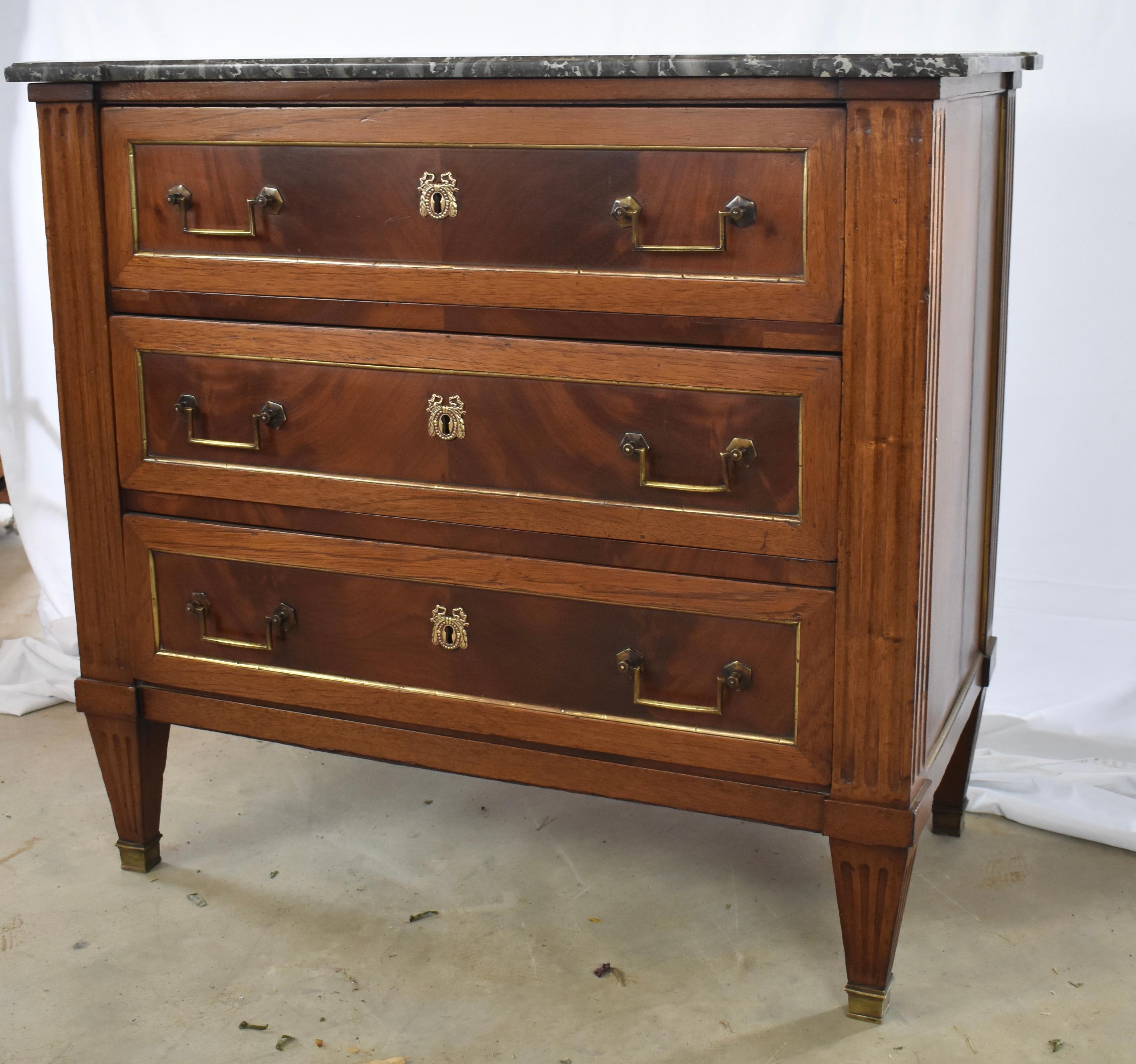 This is an absolutely gorgeous Louis XVI commode built in France in the early 1800s using mahogany and oak for the secondary wood. The chest features three drawers each decorated by book-matched veneer framed by molding and a brass trim. The case