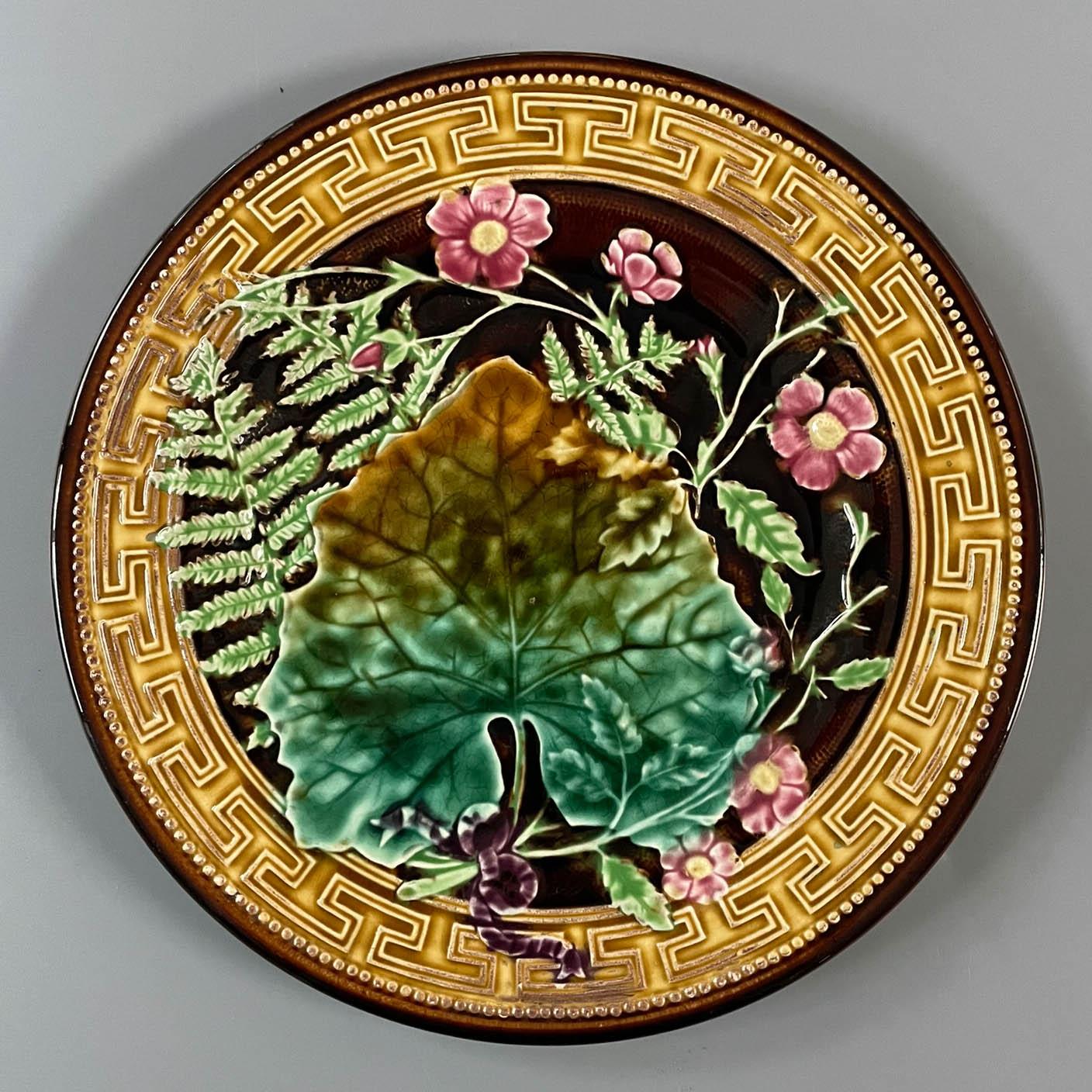 Ein französischer Majolikateller aus glasierter Keramik des 19. Jahrhunderts von H. B. Boulanger Choisy-le-Roi. Muster aus Farnen und rosa Blüten unter einem großen Blatt, gebunden mit einer lila Schleife. Kräftige Grün-, Gelb- und Rosatöne auf