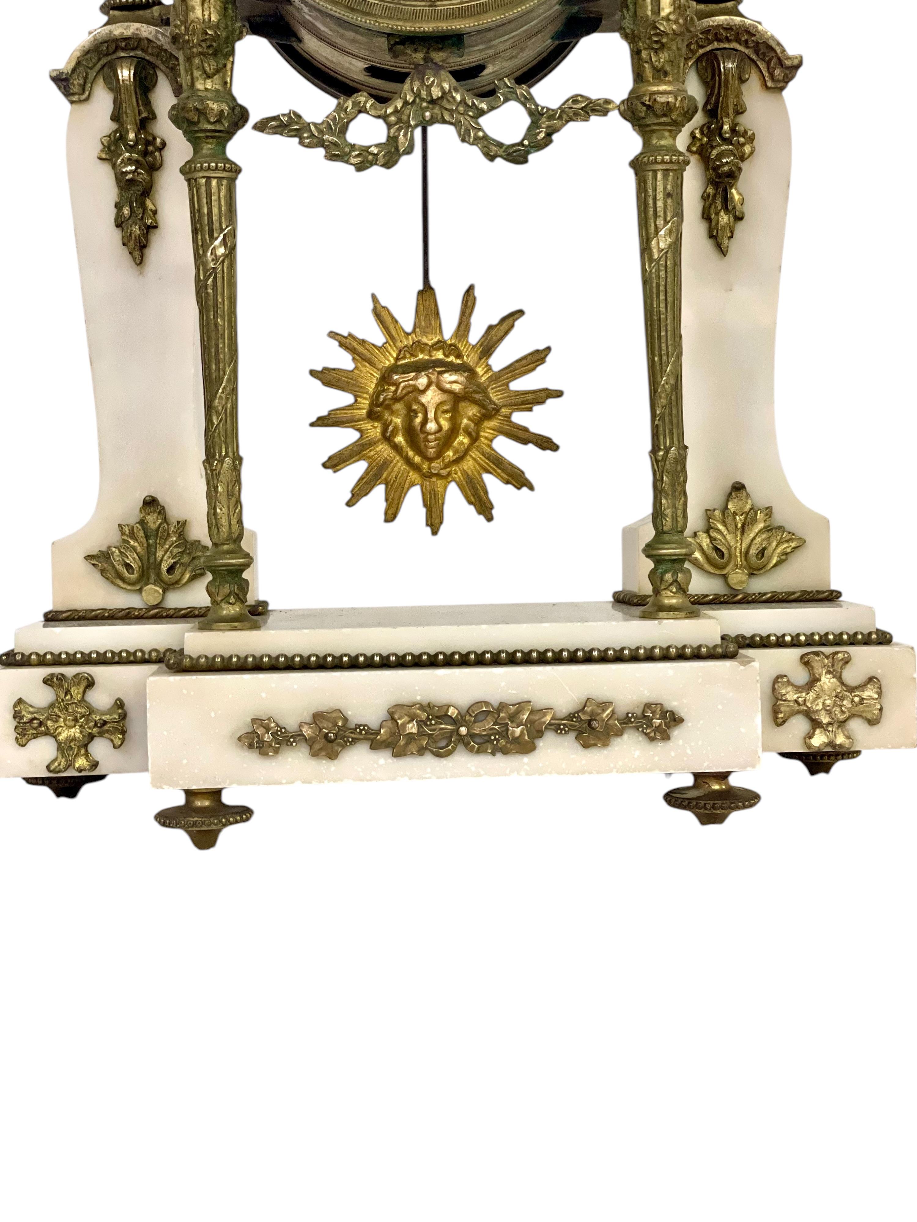 This elegant 19th-century French mantel clock is a refined example of Neoclassical design. Crafted from white marble with exquisite gilt bronze accents, it features intricate details, including a sunburst pendulum, delicate garlands, and floral