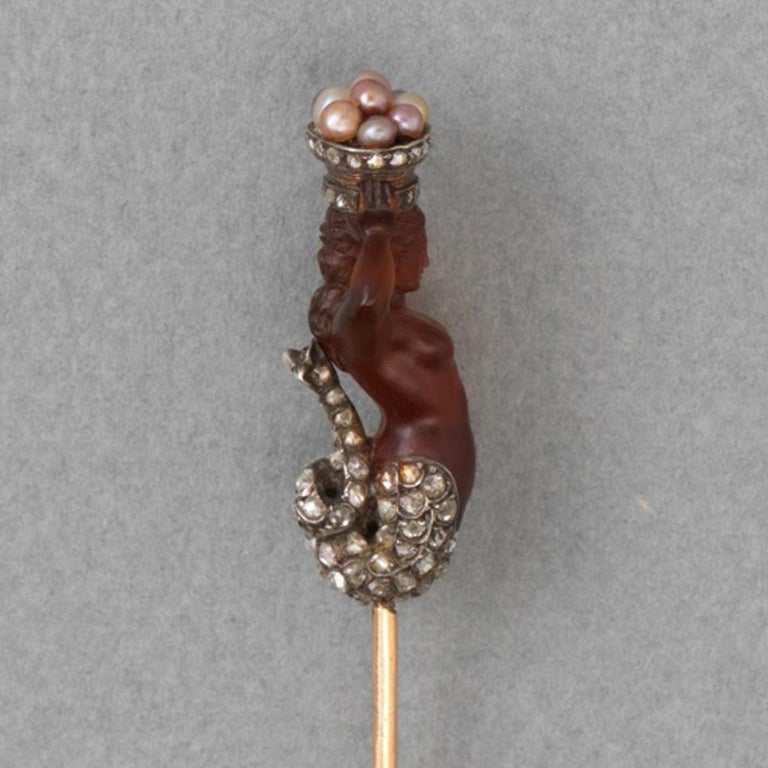A magnificent carved agate mermaid mounted as a stickpin. Her hair, tail, and the edge of the basket are silver and embellished with numerous rose-cut diamonds. Above her head she holds a basket containing different coloured fine pearls, as found in