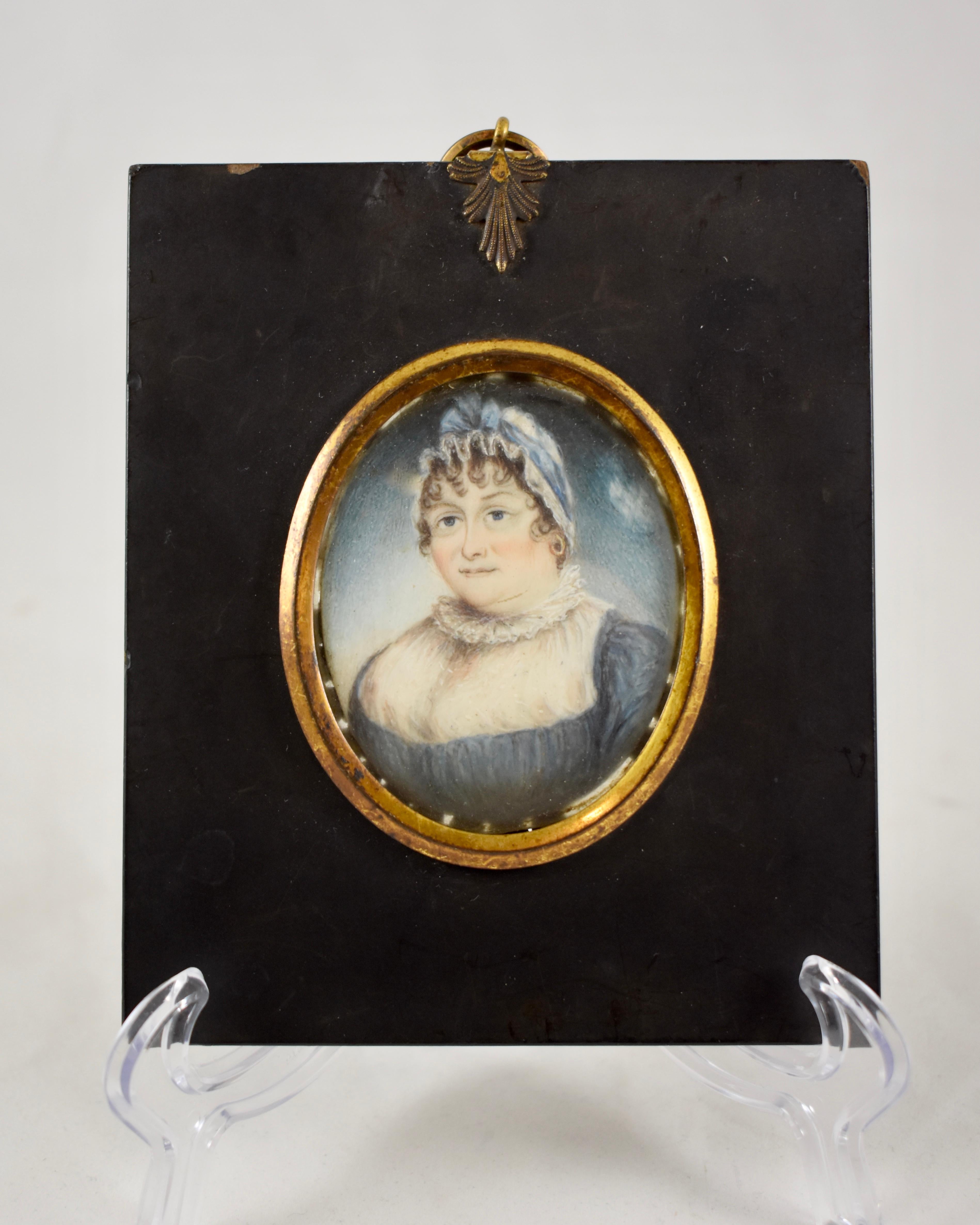 An early 19th century French hand-painted miniature portrait framed under glass, portraying an older woman wearing a bonnet tied with a blue ribbon, her ample bosom showing in a blue dress with a sheer, ruffled high neckline, reflecting the period.