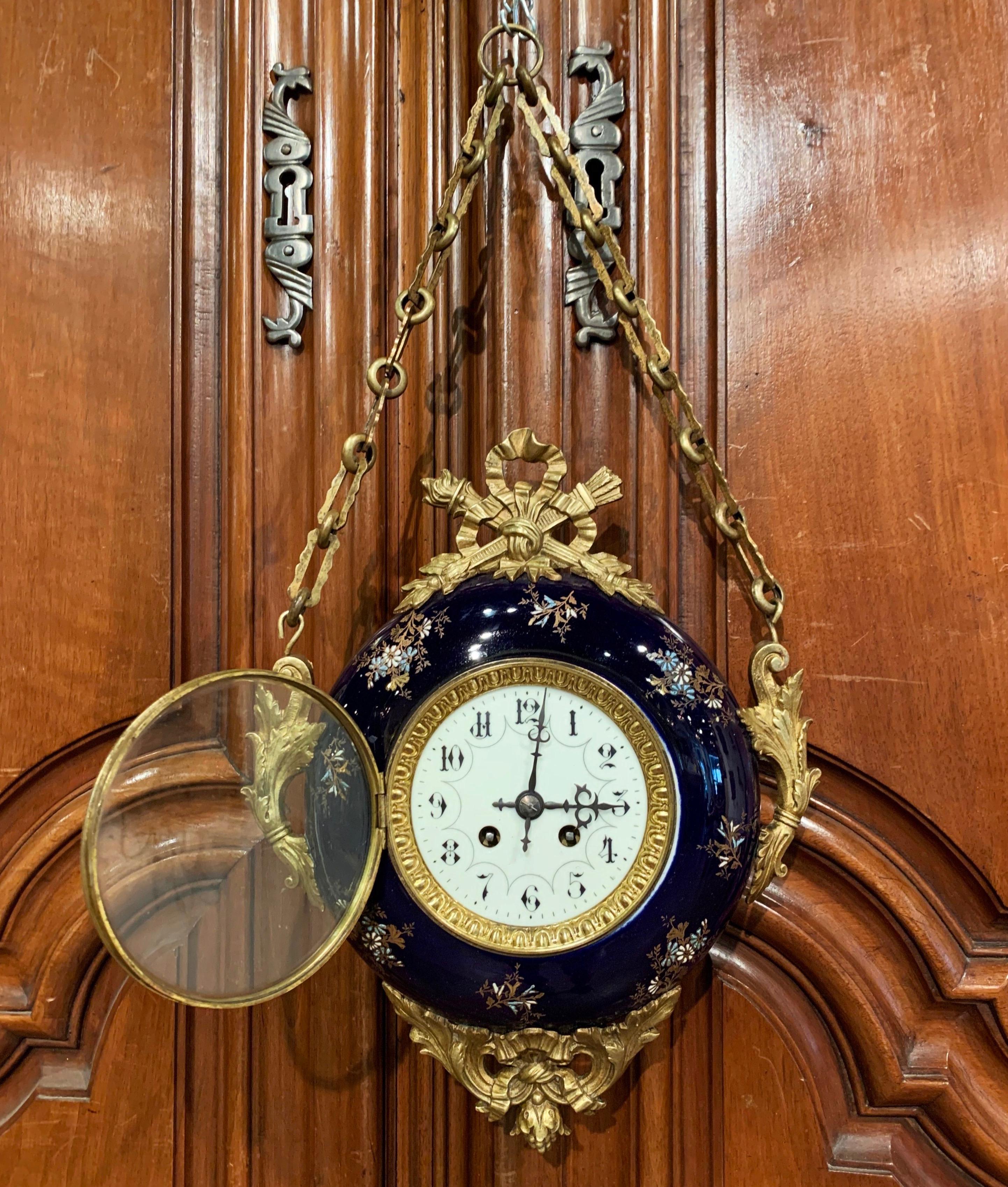 This elegant antique wall hanging clock was crafted in France circa 1870; oval in shape, the colorful clock features brass mounts around the blue cobalt porcelain body decorated with colorful hand painted floral motifs. The central clock face is