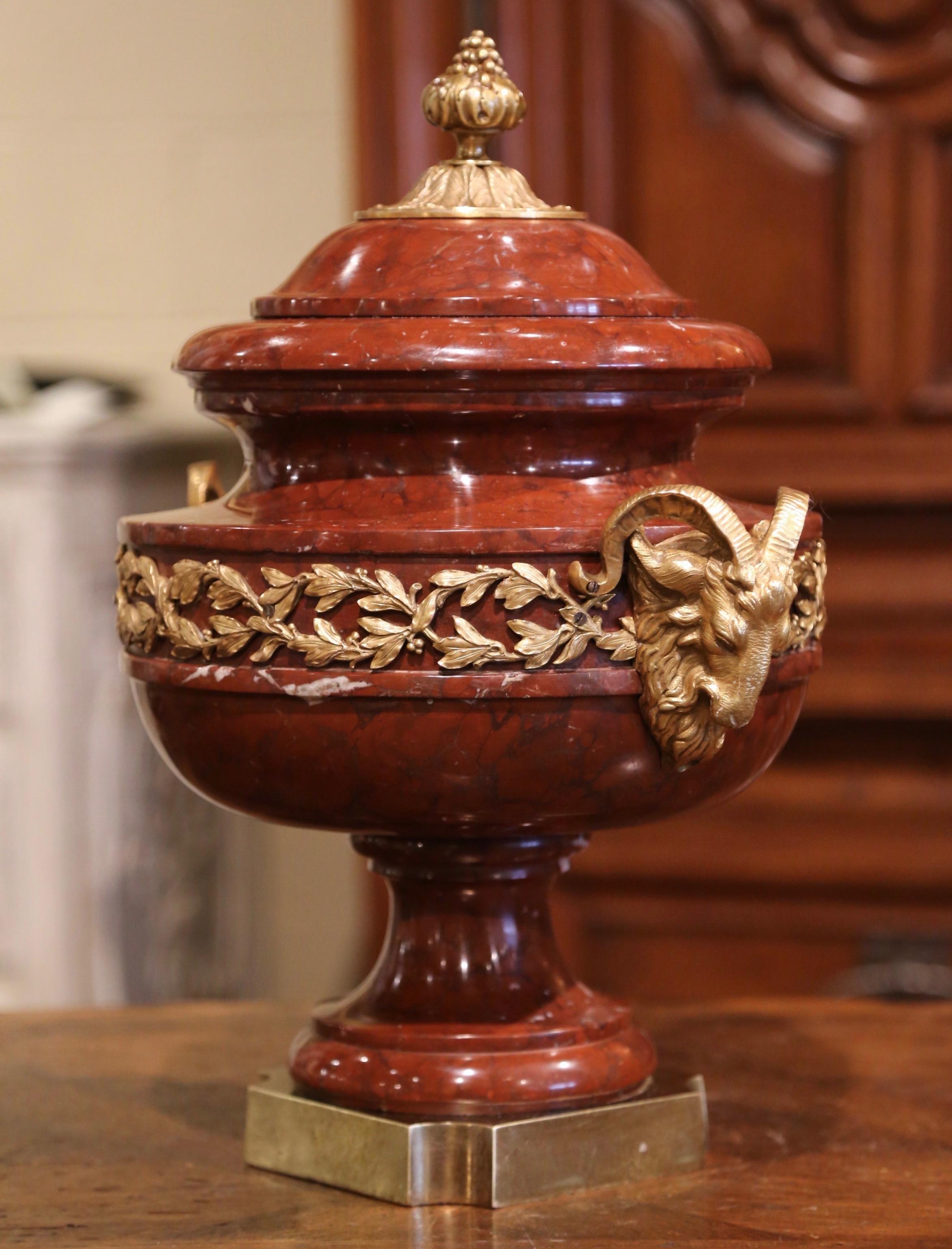Crafted in France, circa 1870 and round in shape, this antique marble urn stands on a square base; the large vessel features a decorative cone-shaped bronze finial at the pediment and is decorated with two bronze side handles with ram figure. The