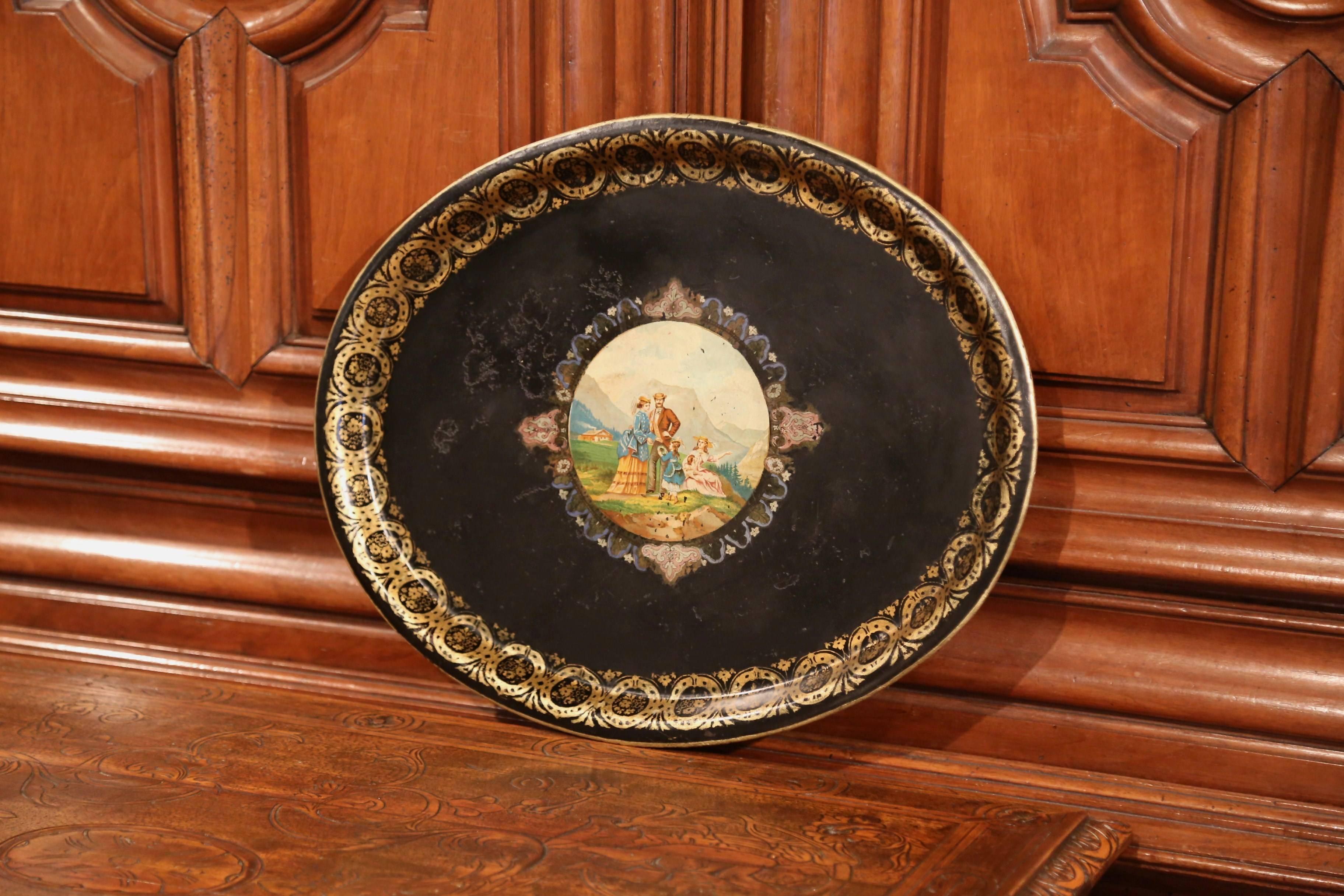 This elegant antique tray was crafted in France, circa 1870; oval in shape, the black tole platter is embellished by hand painted gilt decor around the lip, and features a colorful landscape scene in the center. The scene depicts a family with a