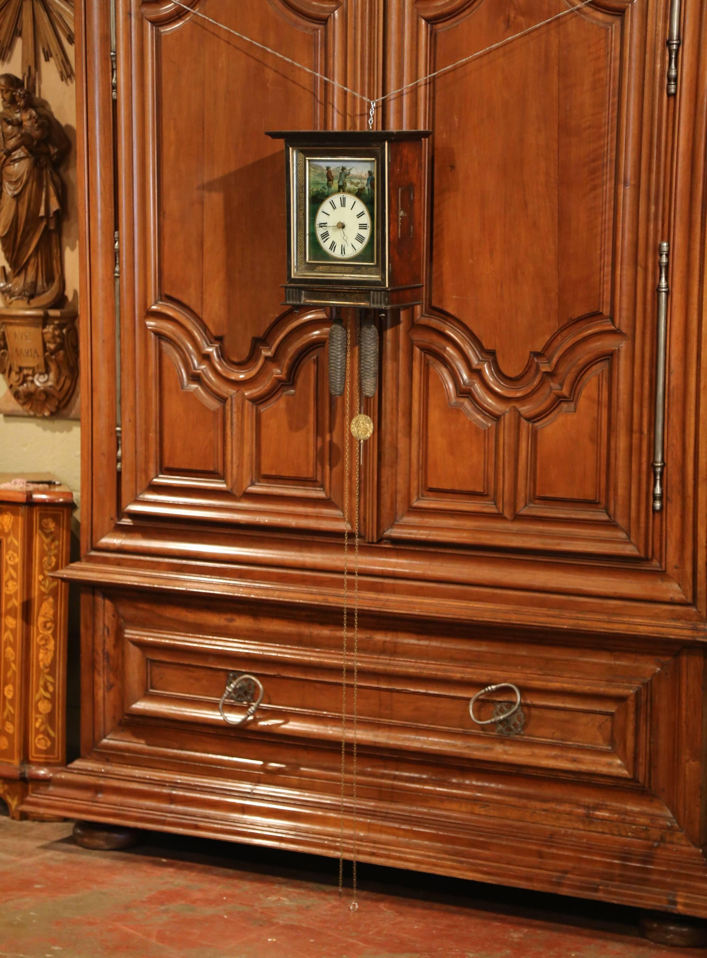 Cette élégante horloge ancienne a été fabriquée en France, vers 1870. Le garde-temps mural noir et doré représente une scène de chasse aux oiseaux peinte à la main en haut, sous la porte vitrée d'origine. On y voit un père qui regarde ses fils