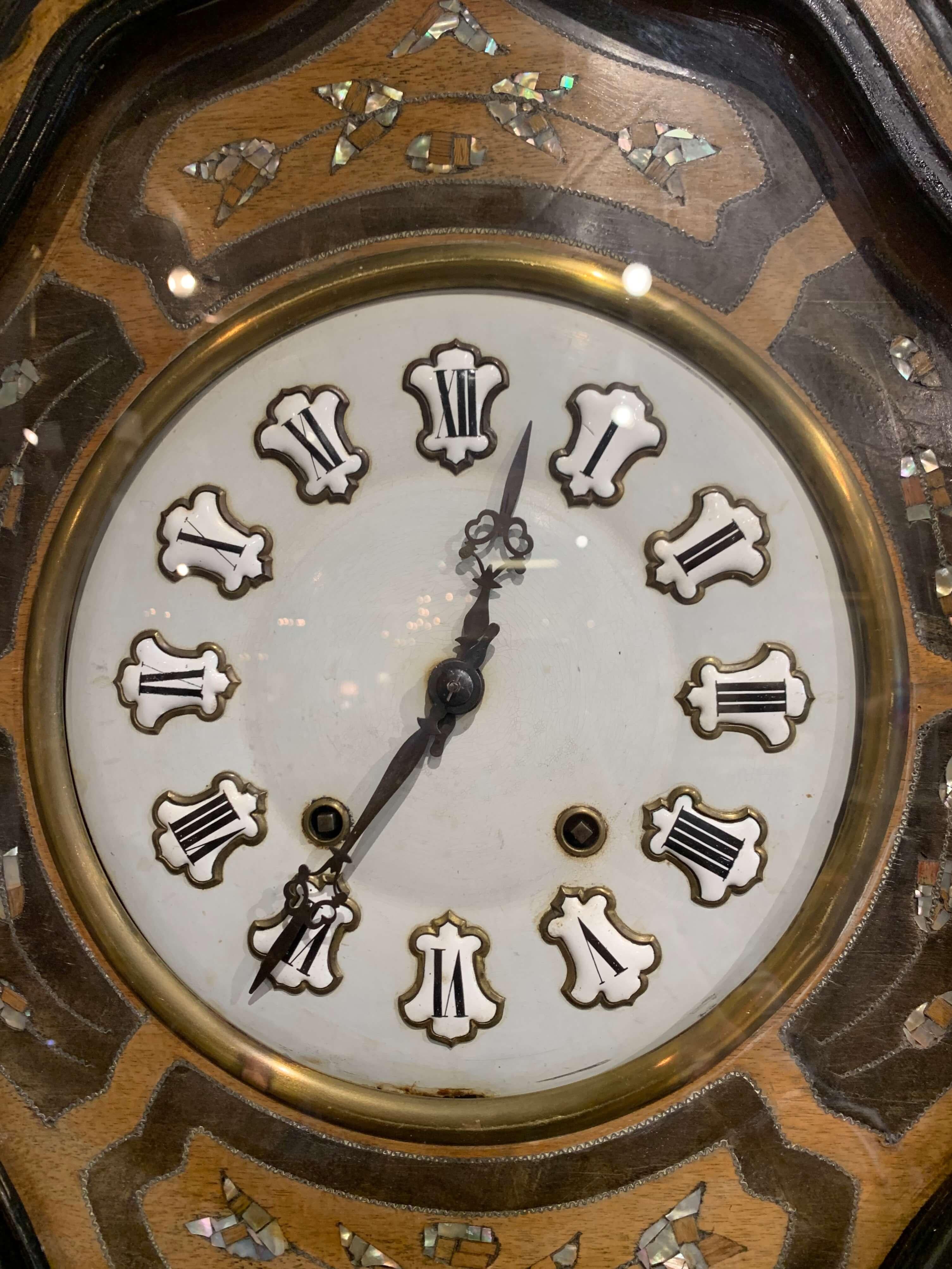 This colorful antique wall hanging clock was crafted in France circa 1870; oval in shape, the time keeper has a two-tone black and wood painted around the perimeter, the time keeper features colorful handcrafted mother of pearl fruit and foliage