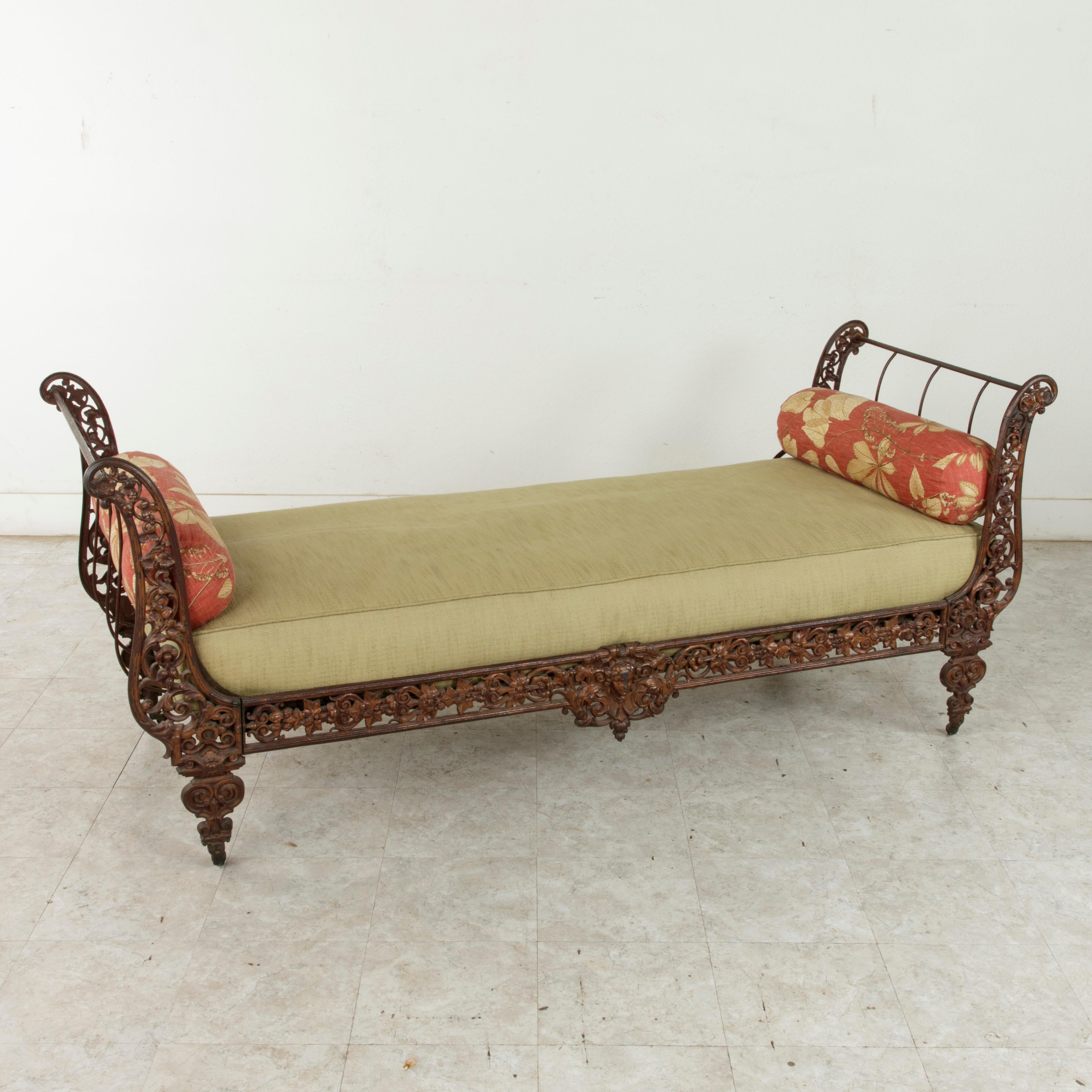 This 19th century French Napoleon III period daybed features a cast iron frame detailed with central cartouche and masque flanked by scrolling branches and leaves that extend out to the curved sides. The frame rests on stylized legs on castors. This