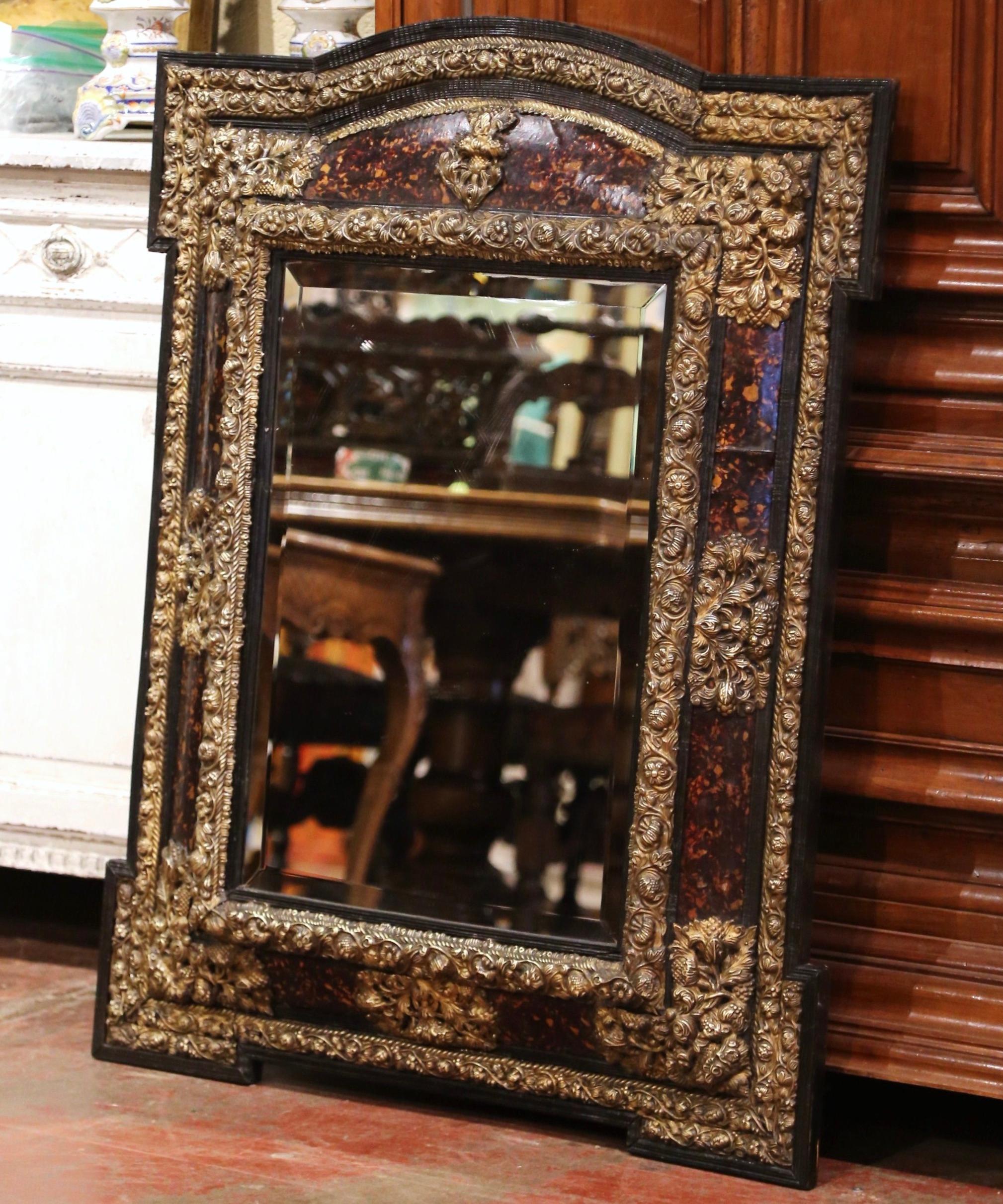 Created in France, circa 1870, the elegant antique wall mirror with overhang corners is rectangular in shape embellished with an arched top. The mirror is decorated with intricate brass repousse floral and fruit motifs in high relief all around the
