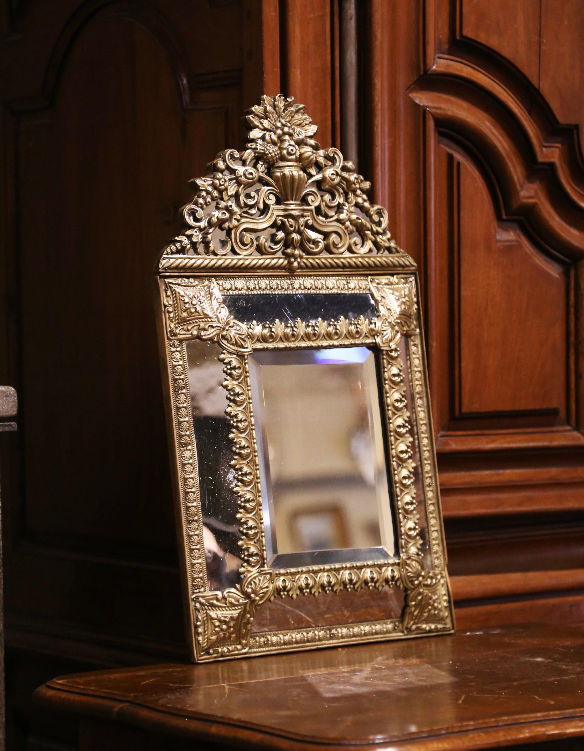 Creata in Francia, intorno al 1870, l'elegante specchiera antica è decorata con intricati motivi a volute in repousse, tra cui un grande cartiglio sul frontone con un cesto di frutta centrale affiancato da volute e fogliame su entrambi i lati. La