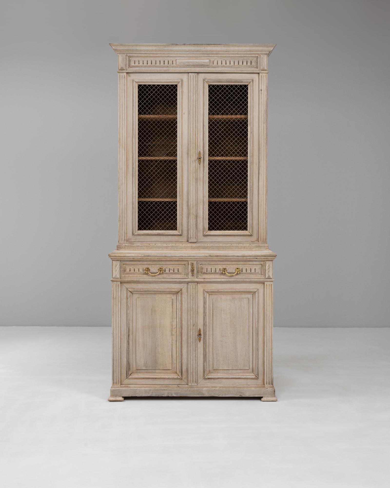 This 19th Century French Neoclassical display cabinet with wire doors exemplifies refined craftsmanship and timeless elegance. The piece is constructed from oak with a softly weathered patina, highlighting its age and authenticity. The upper section