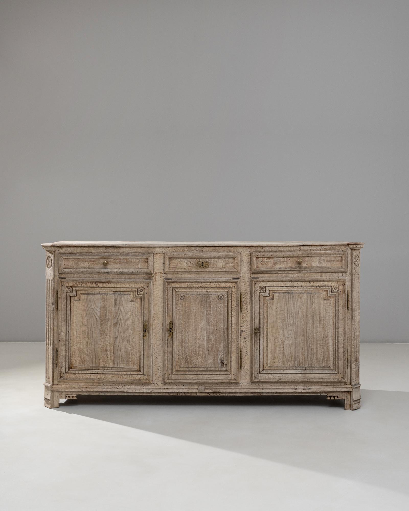 This 19th Century French Neoclassical oak buffet presents a refined balance of symmetry, proportion, and understated ornamentation. Crafted from beautifully grained oak with a softly faded patina, the piece features three paneled doors framed by