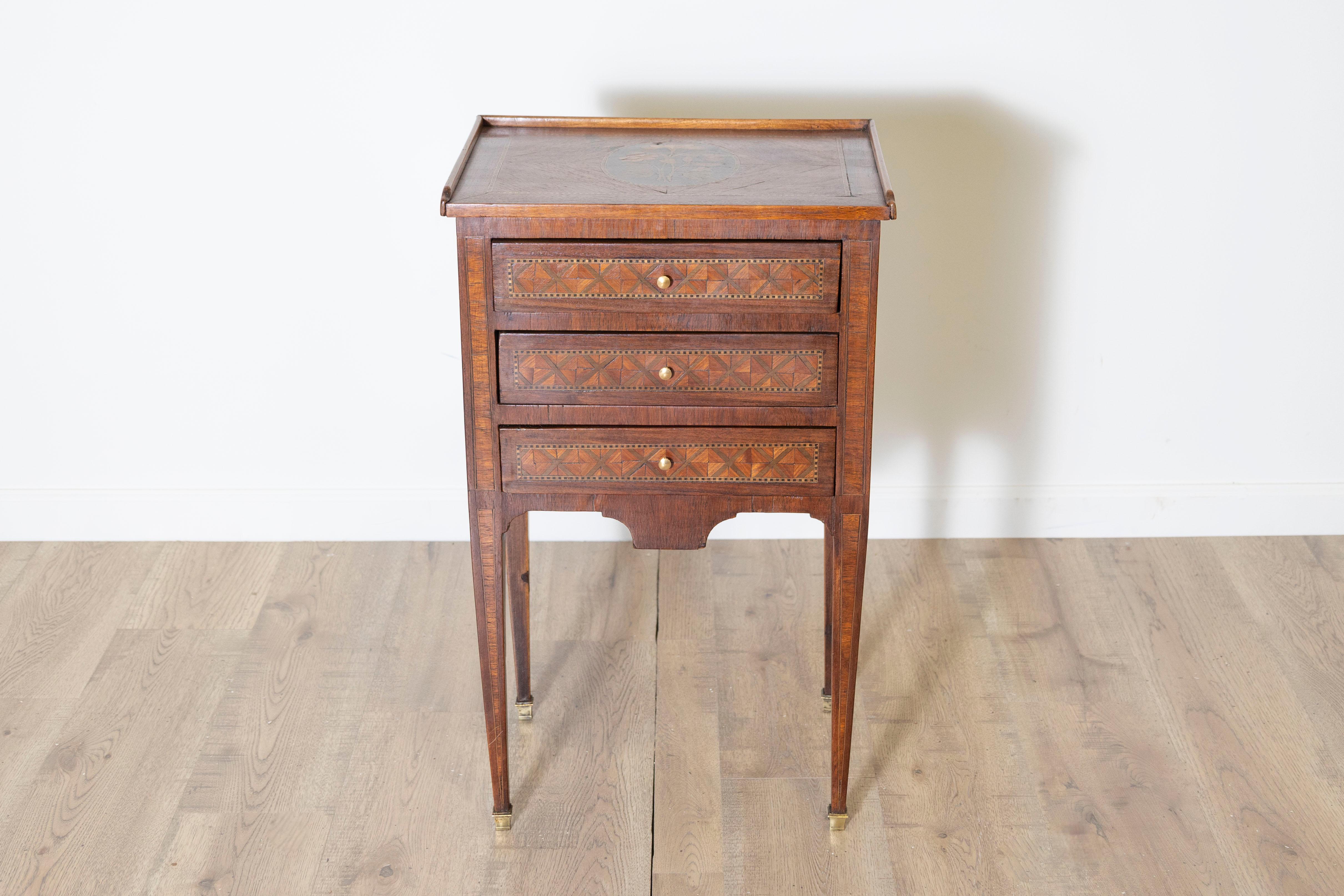 A charming Louis XVI-style bedside table adorned with intricate geometric marquetry and a floral inlay decoration on the top. This exquisite piece showcases the elegance and craftsmanship of the Louis XVI era, combining artistry with functionality.