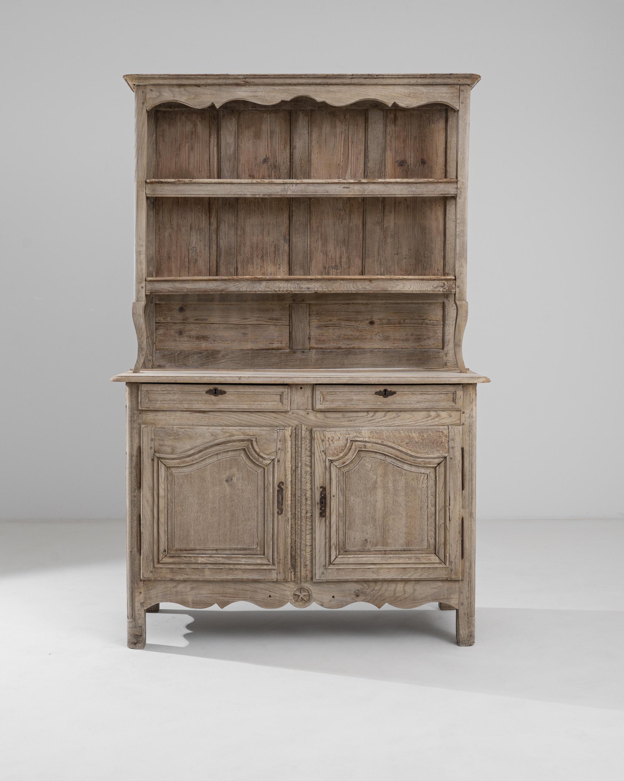 A wooden display cabinet from 19th century France. This neo-classically inspired china cabinet features three upper shelves, a desk surface, two sliding drawers, and lower storage compartments. Pleasant curved scroll patterns are carved along its