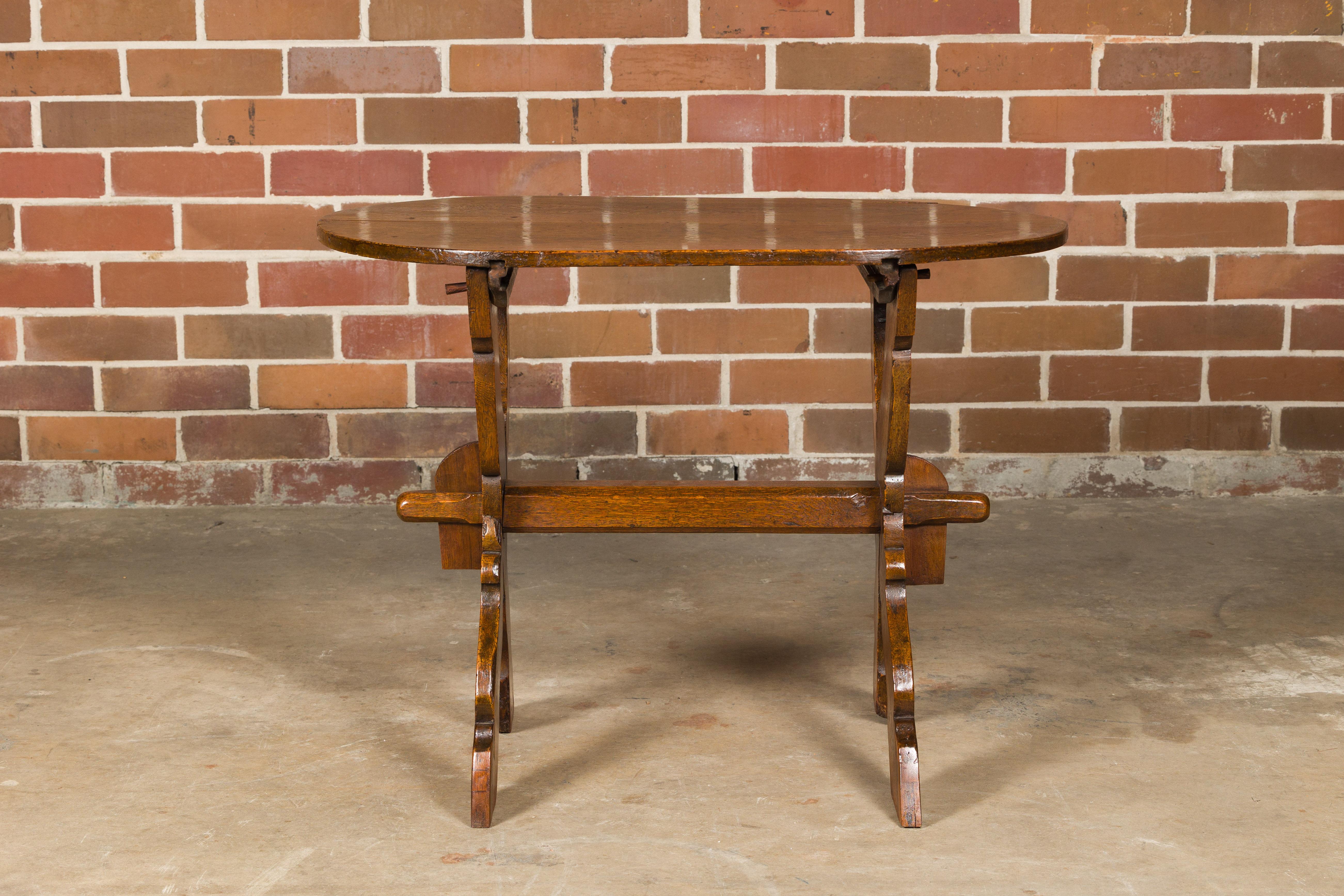 A French oak trestle side table from the 19th century with oval top, double X-Form carved base and cross stretcher. This 19th-century French oak side table exudes rustic elegance with its timeless design and exceptional craftsmanship. The oval top