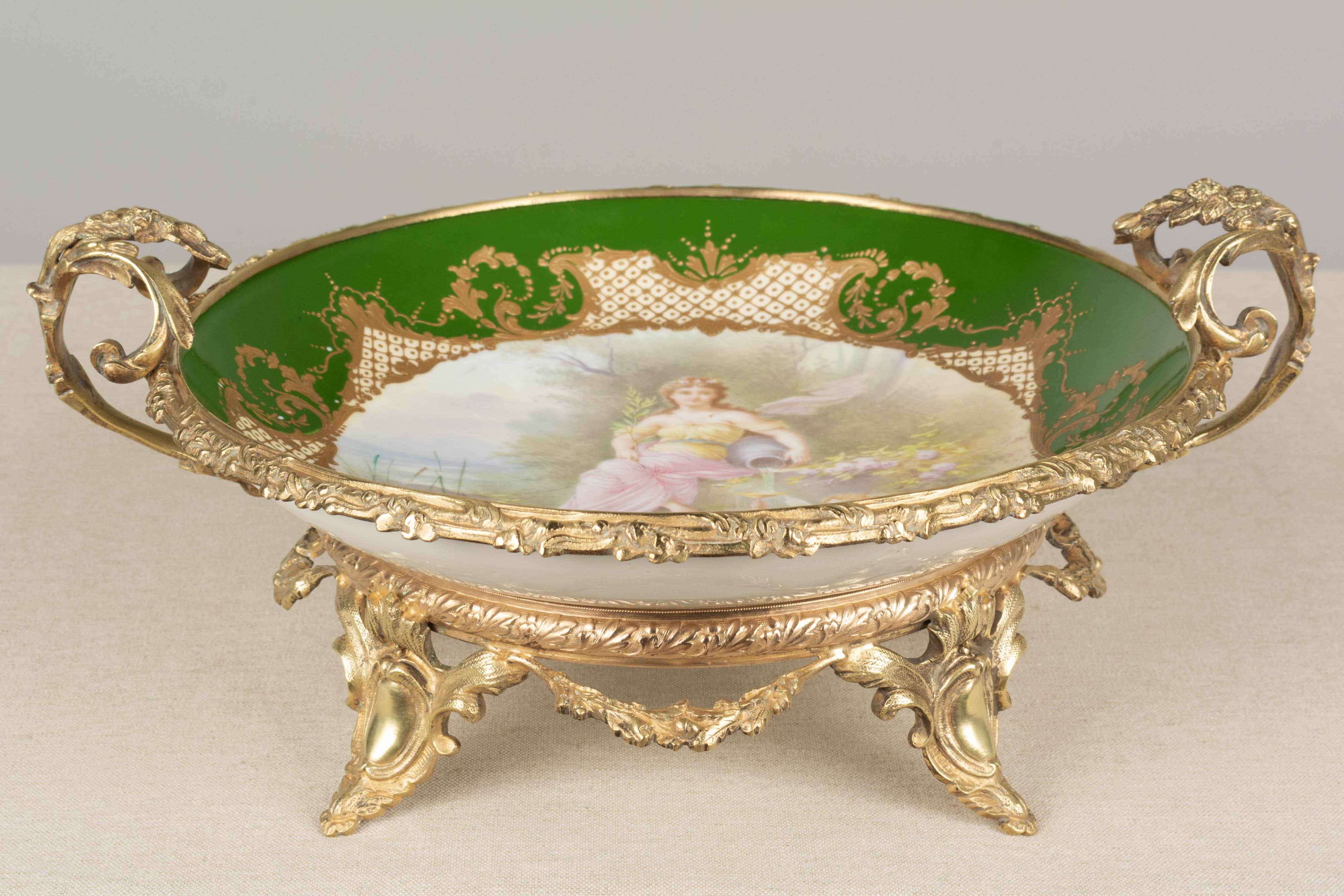 A fine 19th Century French cast ormolu bronze mounted Sèvres porcelain centerpiece with double handles and raised on a footed base. Inset Neo-classical style hand-painted porcelain shallow bowl signed A. Collot with realist Rococo composition of a