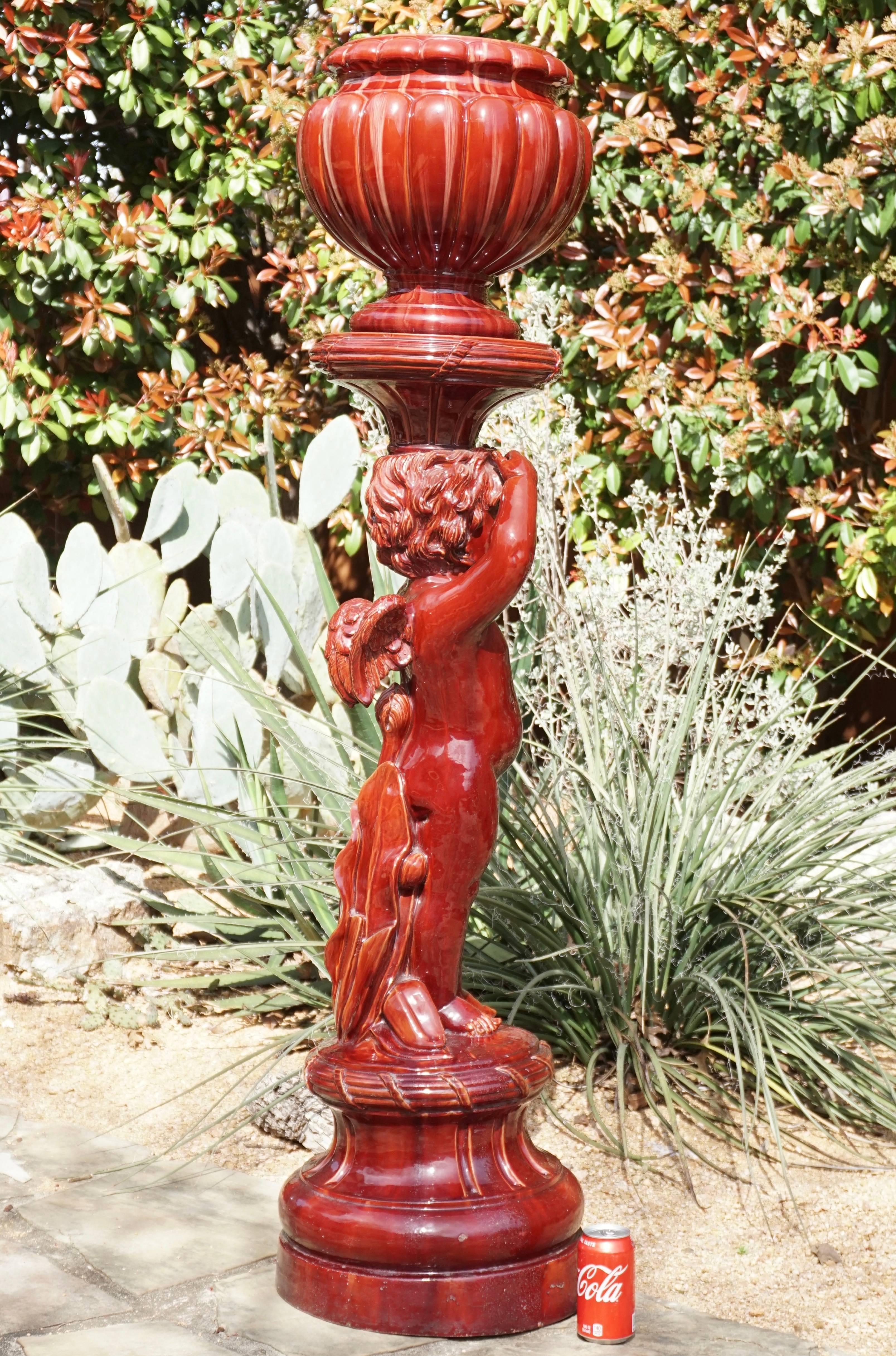 A tall circa 1900 or earlier glazed redware ox blood Majolica jardinière and pedestal representing a winged Putti cherub with Art Nouveau poppy design on verso. The jardinière and pedestal have striations and beauty attributed to Clement Massier