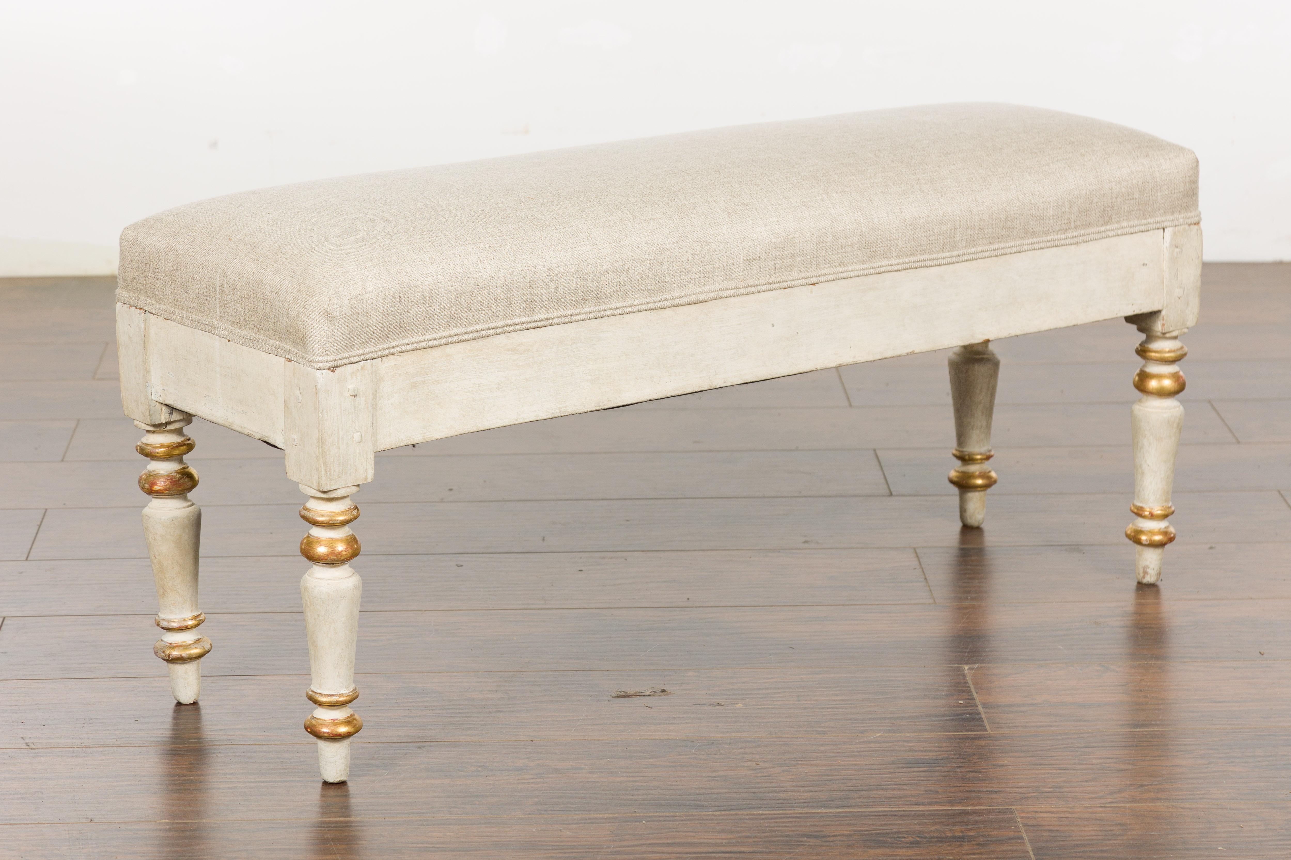 A French painted wood bench from the 19th century with gilt accentuation, turned legs and new linen upholstery. This exquisite French painted wood bench from the 19th century exudes classic elegance, bringing a touch of historic charm to any space.