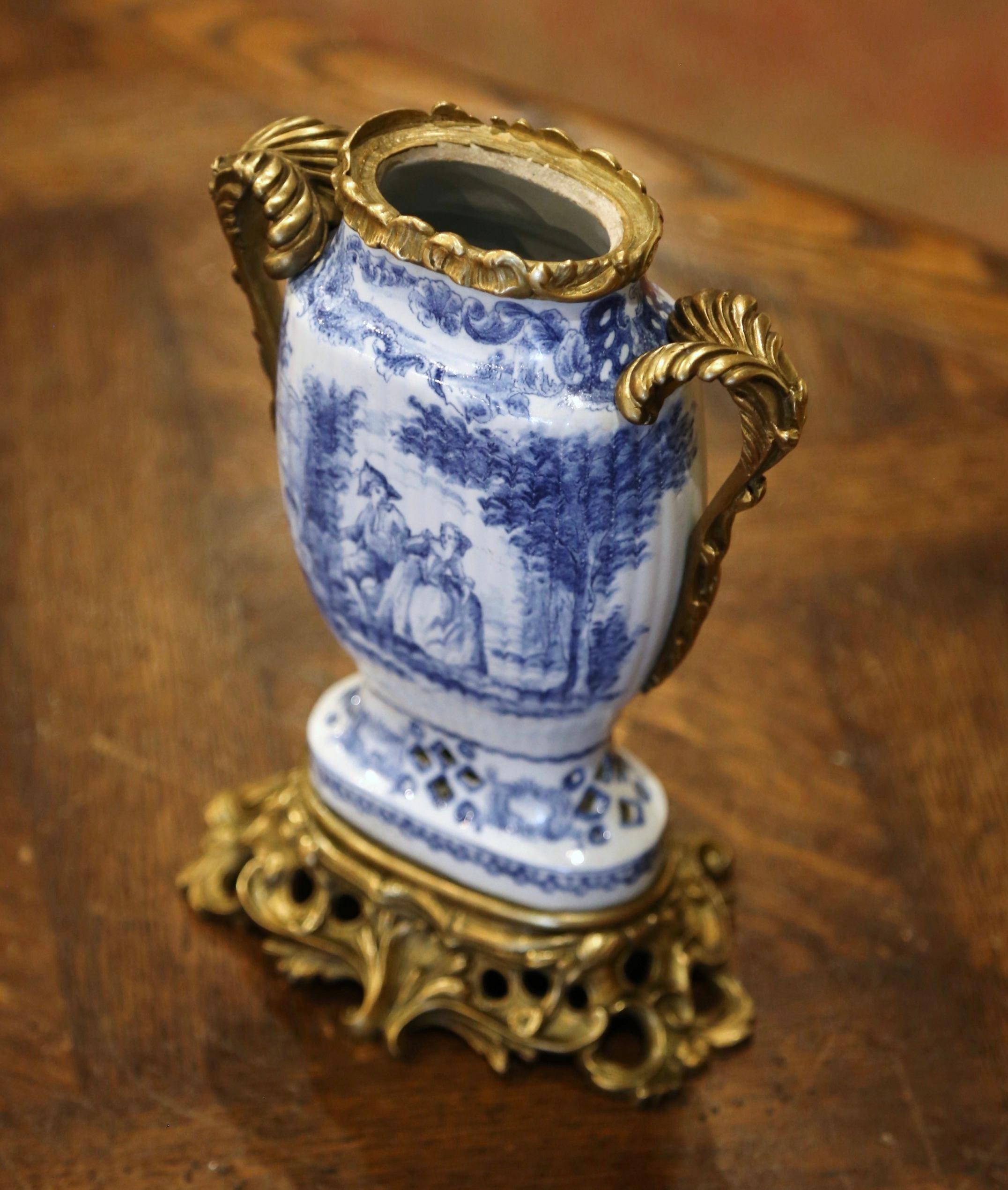 Crafted in Holland, circa 1870, the antique faience vase stands on an intricate bronze stand dressed with two decorative handles and embellished with a bronze rim. The vase is decorated with a hand painted blue and white courting scene medallion on