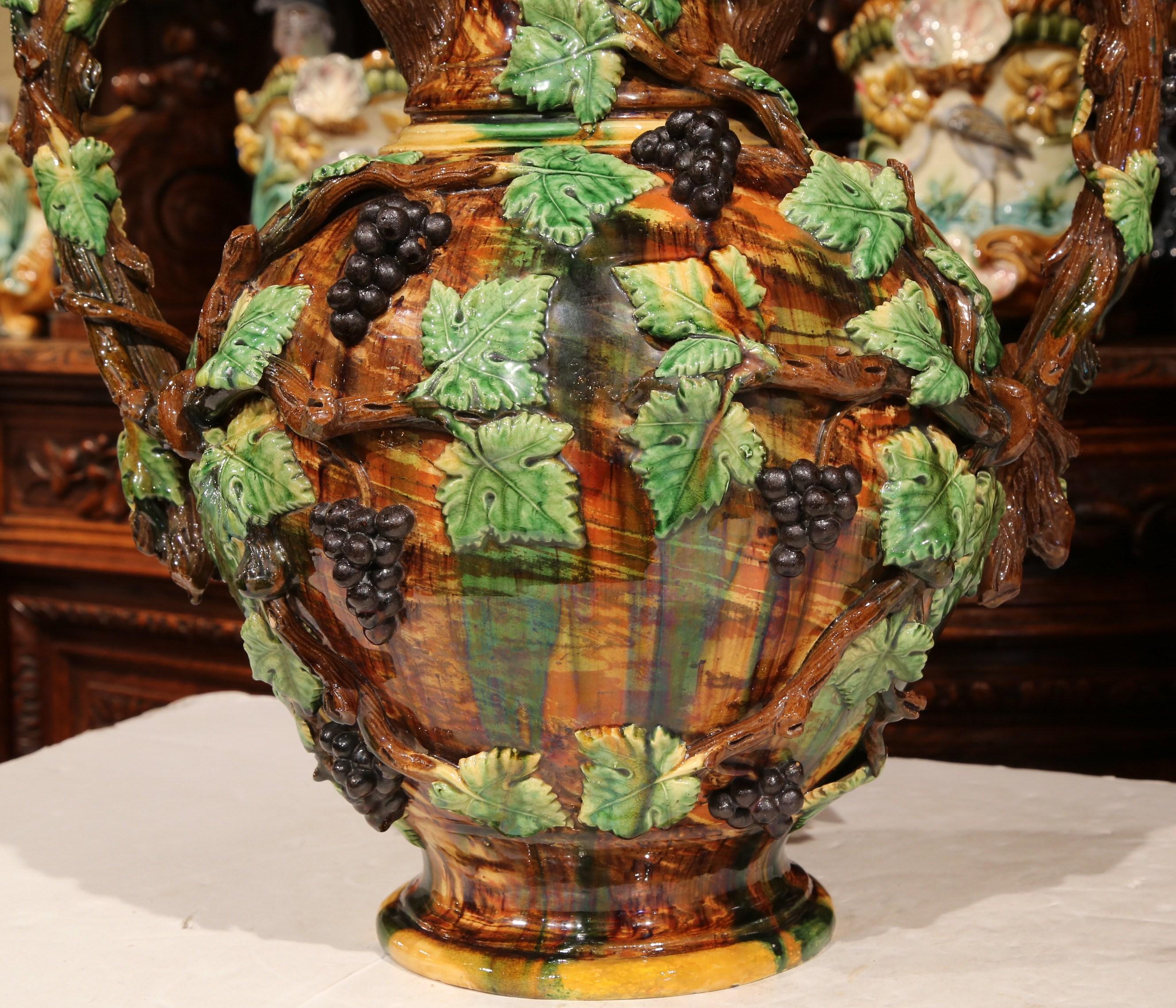 This large, antique hand painted ceramic wine jar was created in France, circa 1890. The tall vase with brown handles shaped like old vines, is embellished with colorful black grapes and green cabernet leaves in high relief. This colorful vessel is