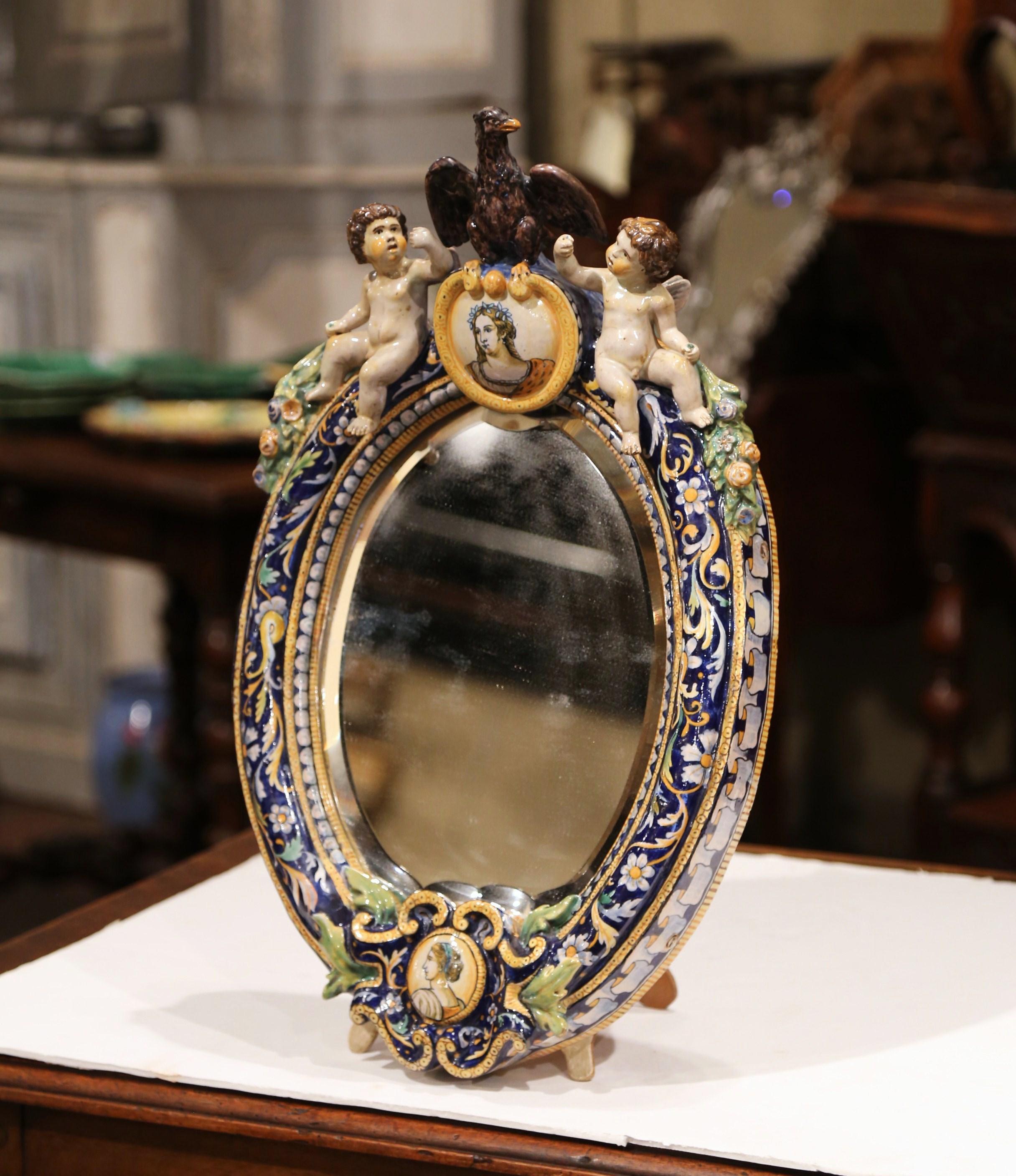 Decorate a counter with this elegant freestanding ceramic table mirror. Crafted in France, circa 1860, the colorful mirror features an eagle figure at the pediment sited over a woman face medallion, and flanked by two cherubs on the sides decorated