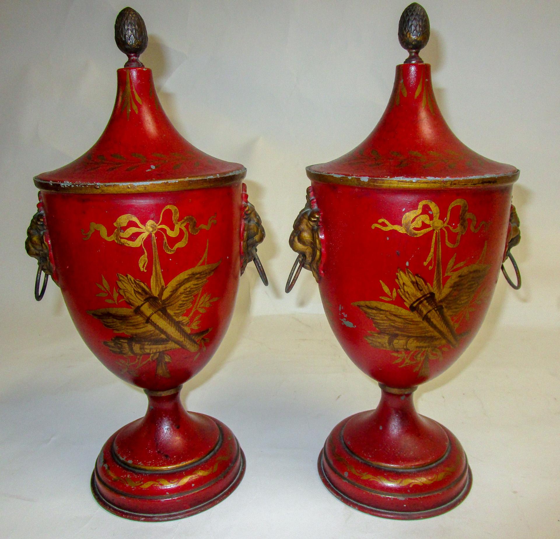 This very handsome pair of French toleware lidded urns, in a deep red color with gold gilt accents, feature lion head handles on either side and artichoke finials atop the lids. Each urn is intricately hand painted with a different scene of