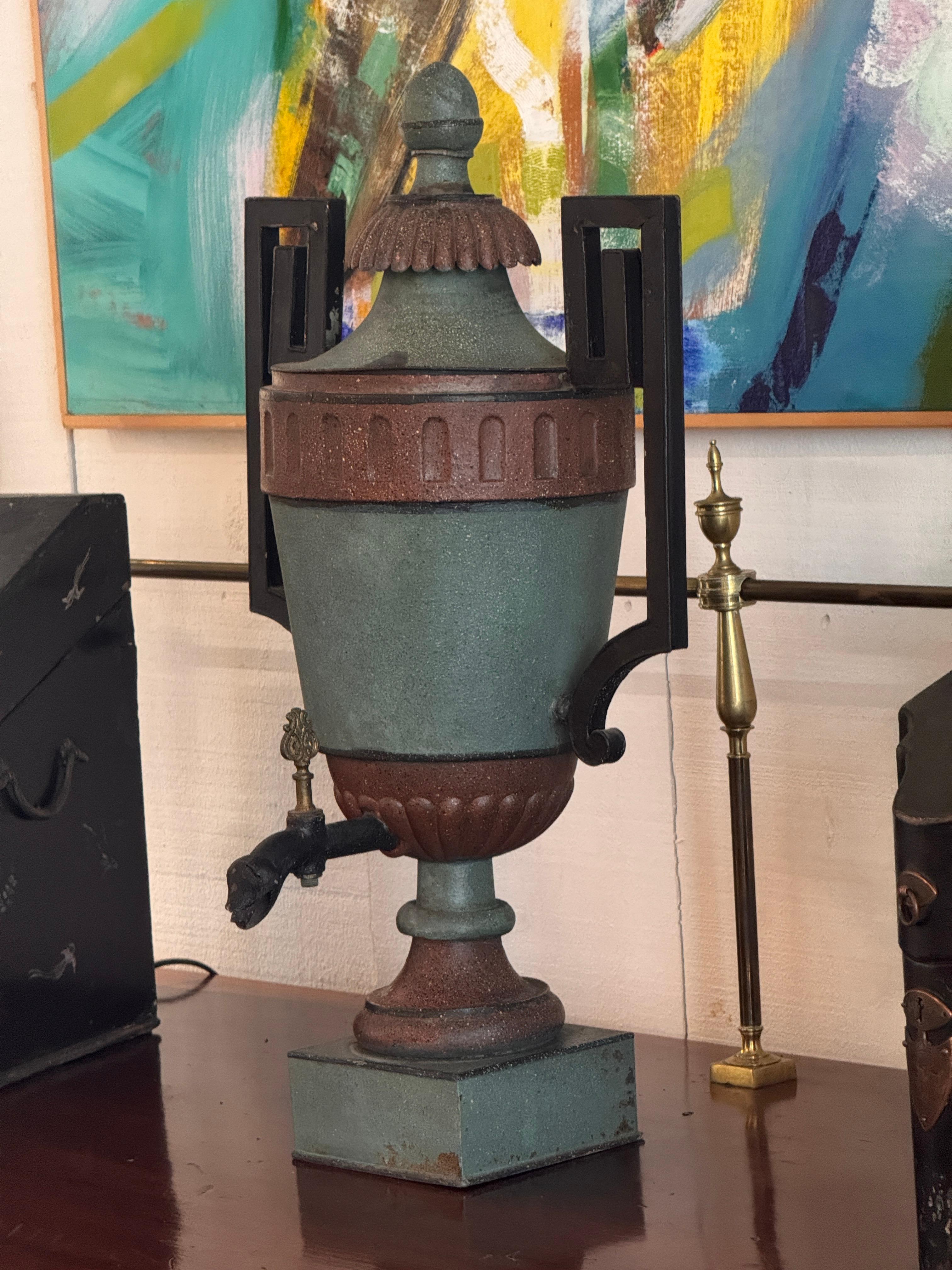 Painted tole urn with a fluted lower body, square plinth base, and rectangular open side handles. The spout is cast in an animal-head form with a small brass turn key. Surface shows worn painted layers consistent with tole work, as seen in the