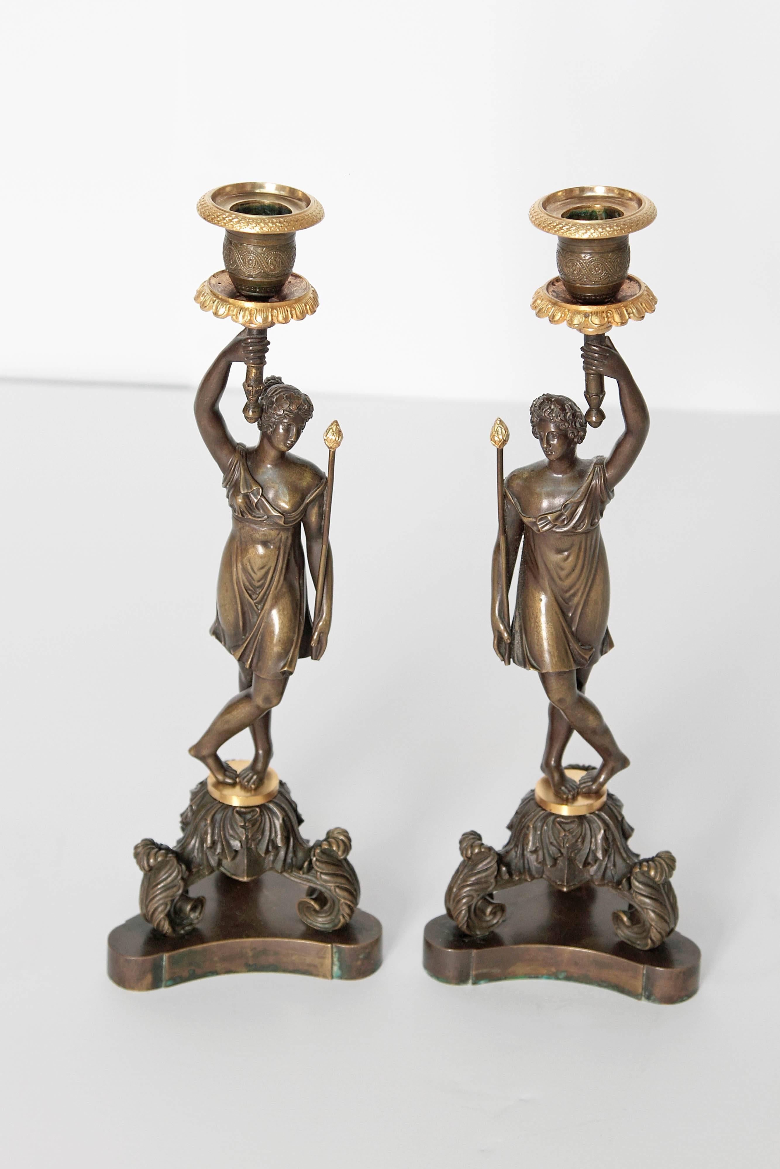 A pair of bronze and gilt bronze candlesticks of well-cast classical women holding a torch which holds the candle. They stand on a triangular plinth base and are supported with tripartite acanthus leaf volutes, 19th century, France.