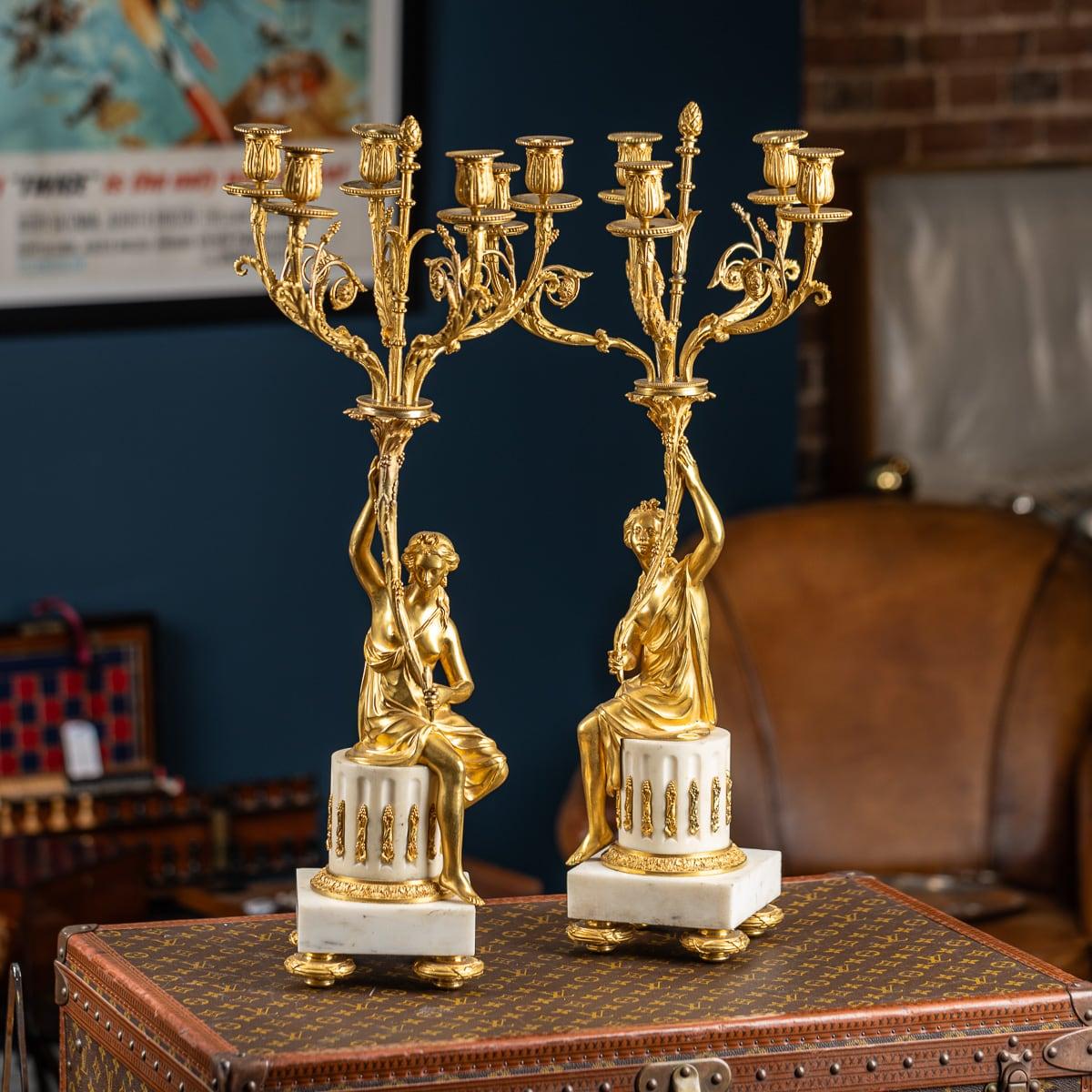 Antique French del XIX secolo, coppia di candelabri a cinque luci in bronzo dorato e marmo bianco in stile Luigi XVI. Ognuno di essi è caratterizzato da bracci a volute foliate che si estendono da un gambo centrale con una ghianda finale, sostenuto