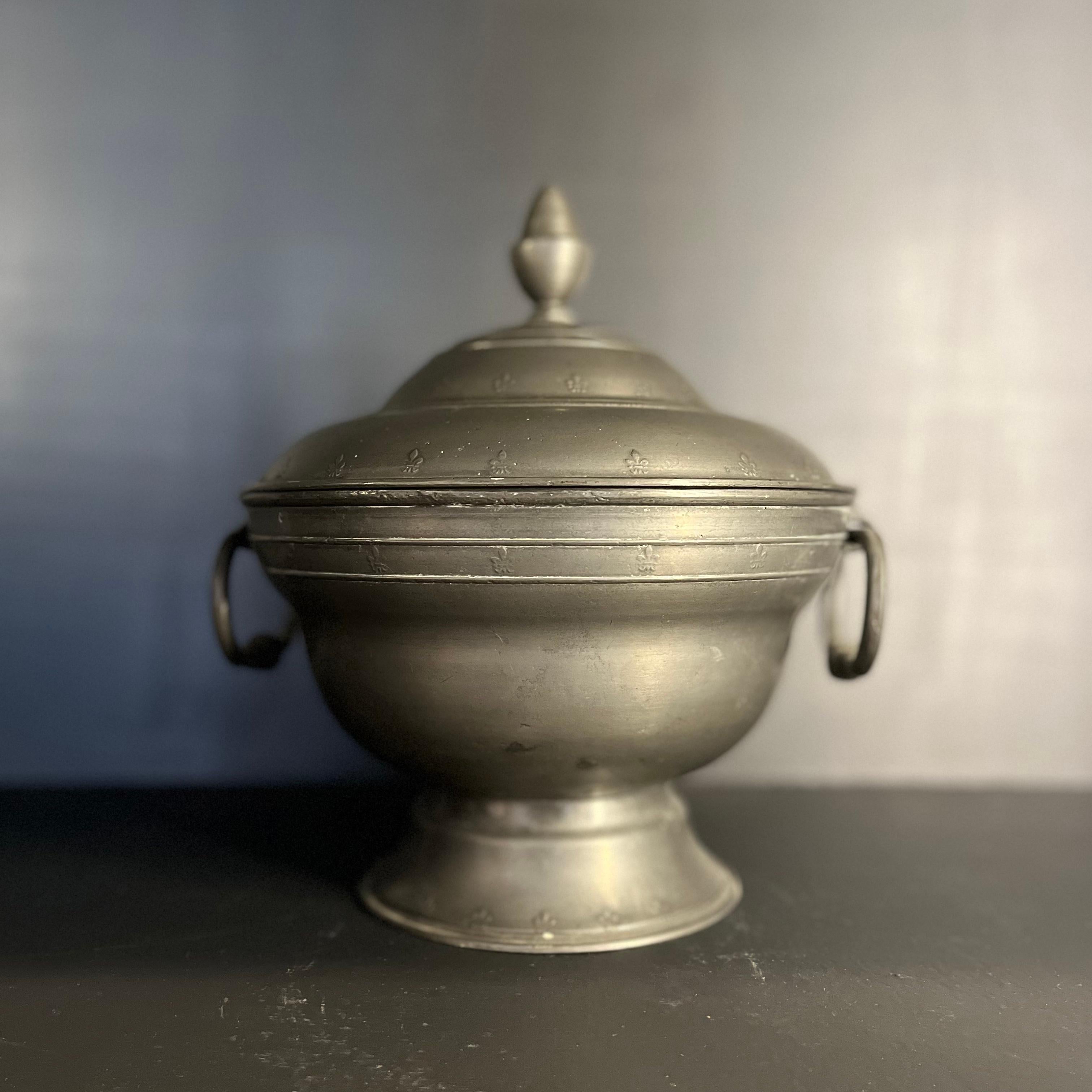 Mid-19th century round pewter soup tureen from France. Featuring an elevated base, two ring-shaped carrying handles, lid with almandine finial topper and delicate decorative raised fleur de lis accenting. Rustic yet refined, the piece could work in