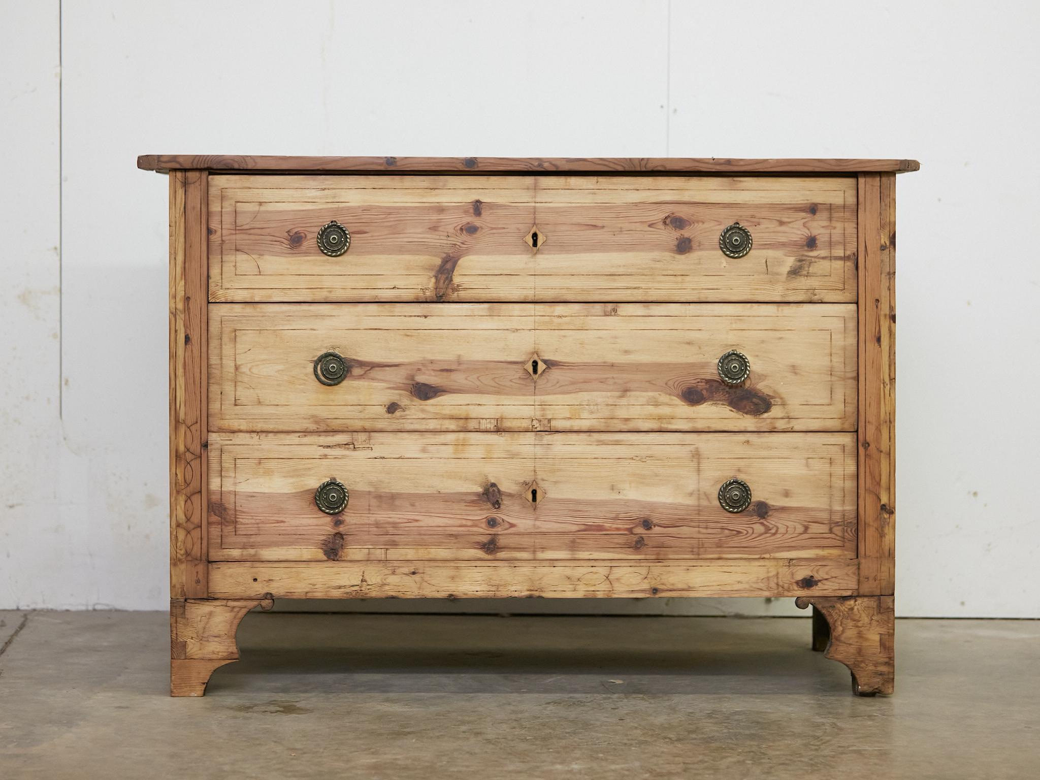 A French pine commode from the 19th century with three drawers, incised motifs, carved feet and nicely weathered appearance. This 19th-century French pine commode exudes an enchanting rustic flair, blending timeless craftsmanship with warm, lived-in