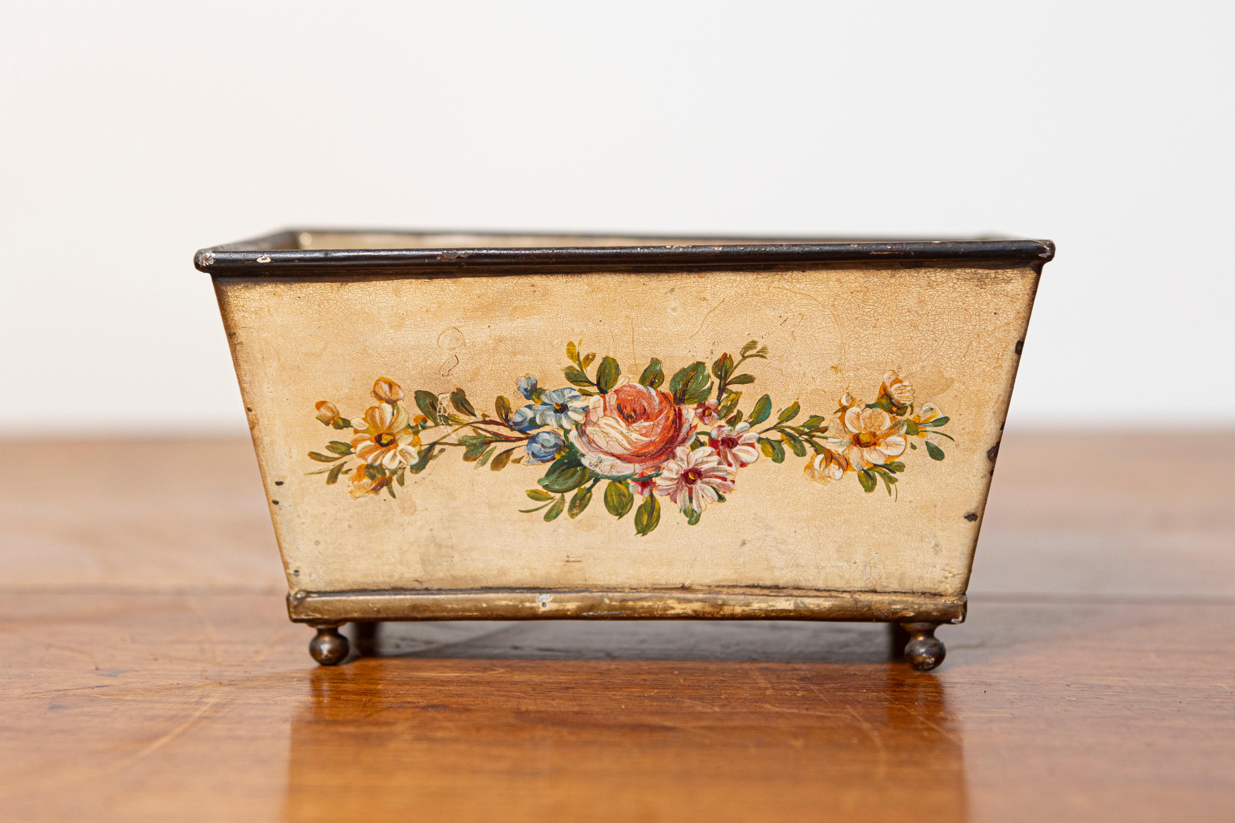 Questa piccola fioriera francese del XIX secolo è realizzata in lamiera verniciata e presenta delicati disegni floreali dipinti a mano su entrambi i lati. Su un morbido sfondo beige, la fioriera mette in mostra rose e altri fiori vivaci, aggiungendo