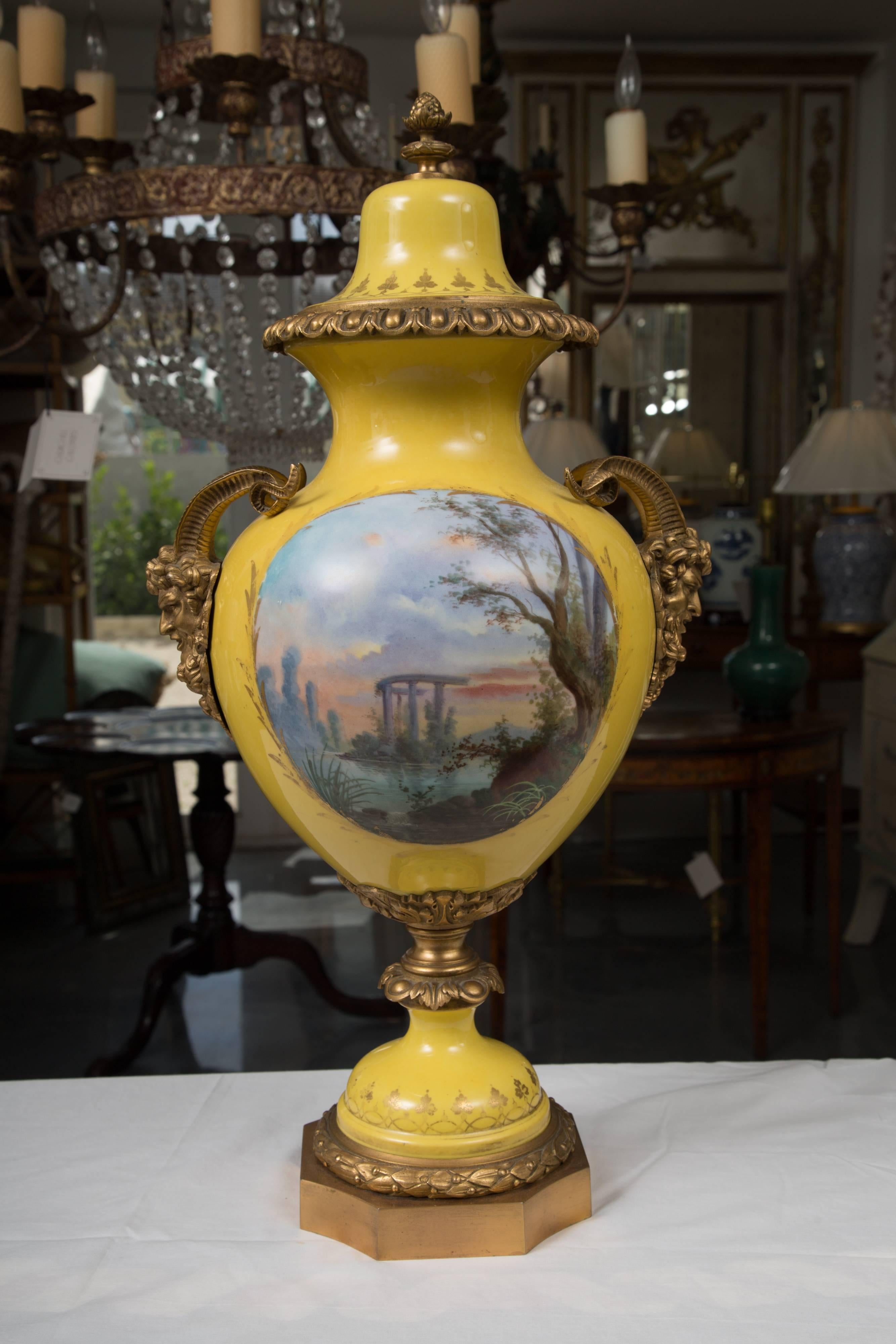 This soft yellow hand-painted porcelain large-scale urn depicts lovers with cherubs in background. The reverse side shows a classic naturalistic setting. The body of the urn is decorated with impressive rams heads. There is a removable lid and the
