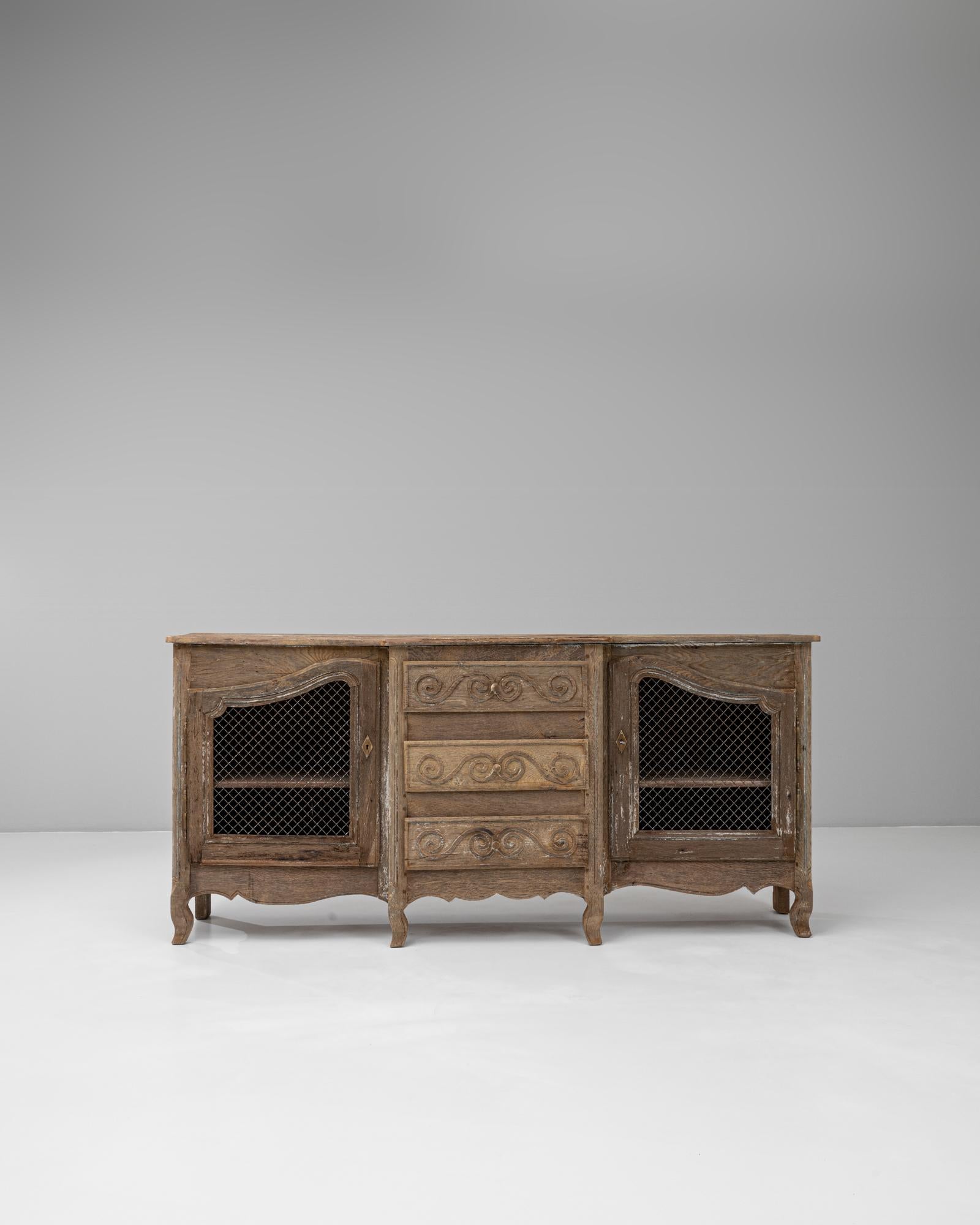 This 19th Century French Provincial buffet beautifully captures the rustic refinement of the period, blending functionality with artisan detail. Crafted in oak with a timeworn patina, it features two side cabinets with wire mesh doors, offering both