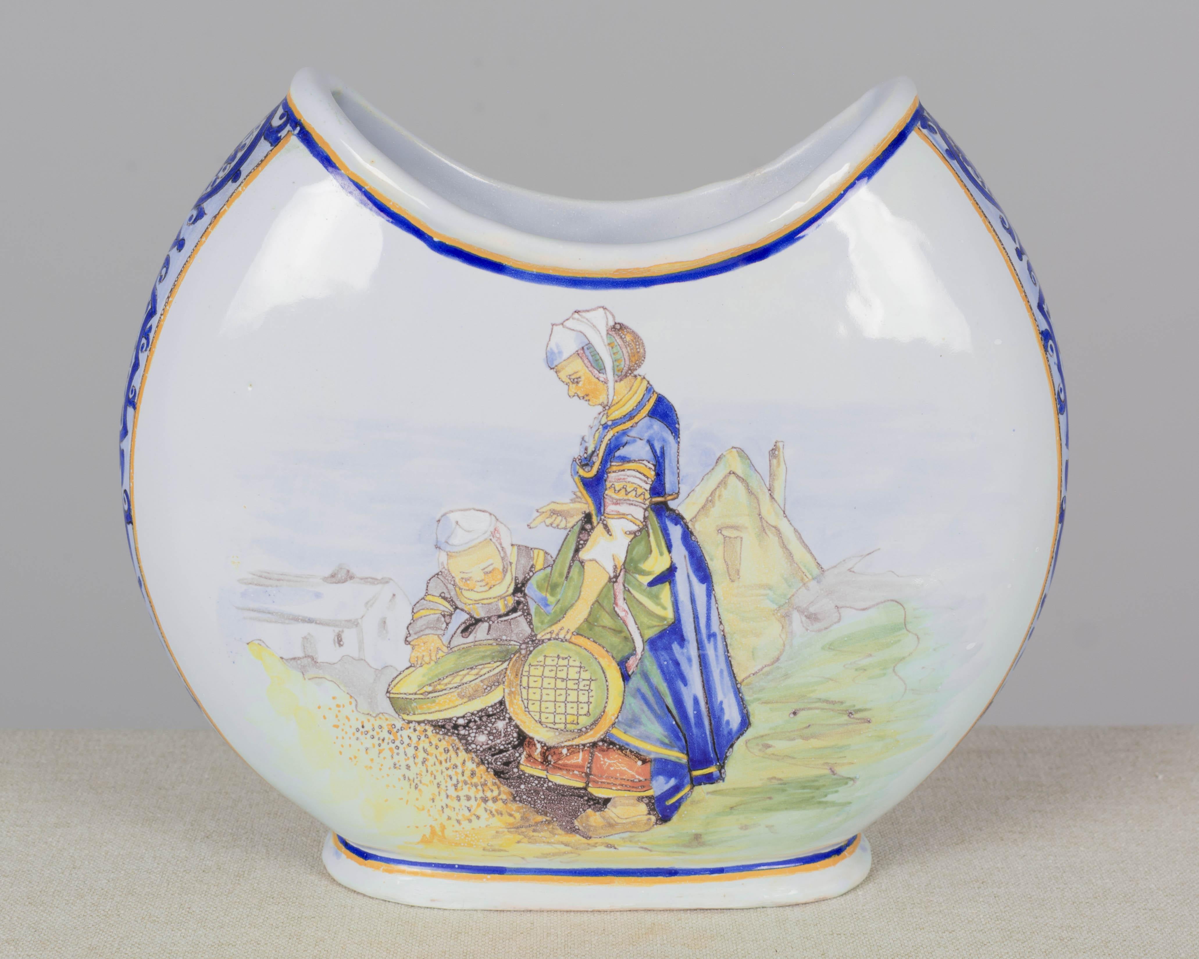 Jarrón francés de loza de Quimper del siglo XIX, obra de Porquier-Beau, pintado a mano con escenas de una madre y una hija bretonas tamizando trigo y un padre y su hijo afilando cuchillos. Bandas de decoración foliada azul oscuro en los laterales.
