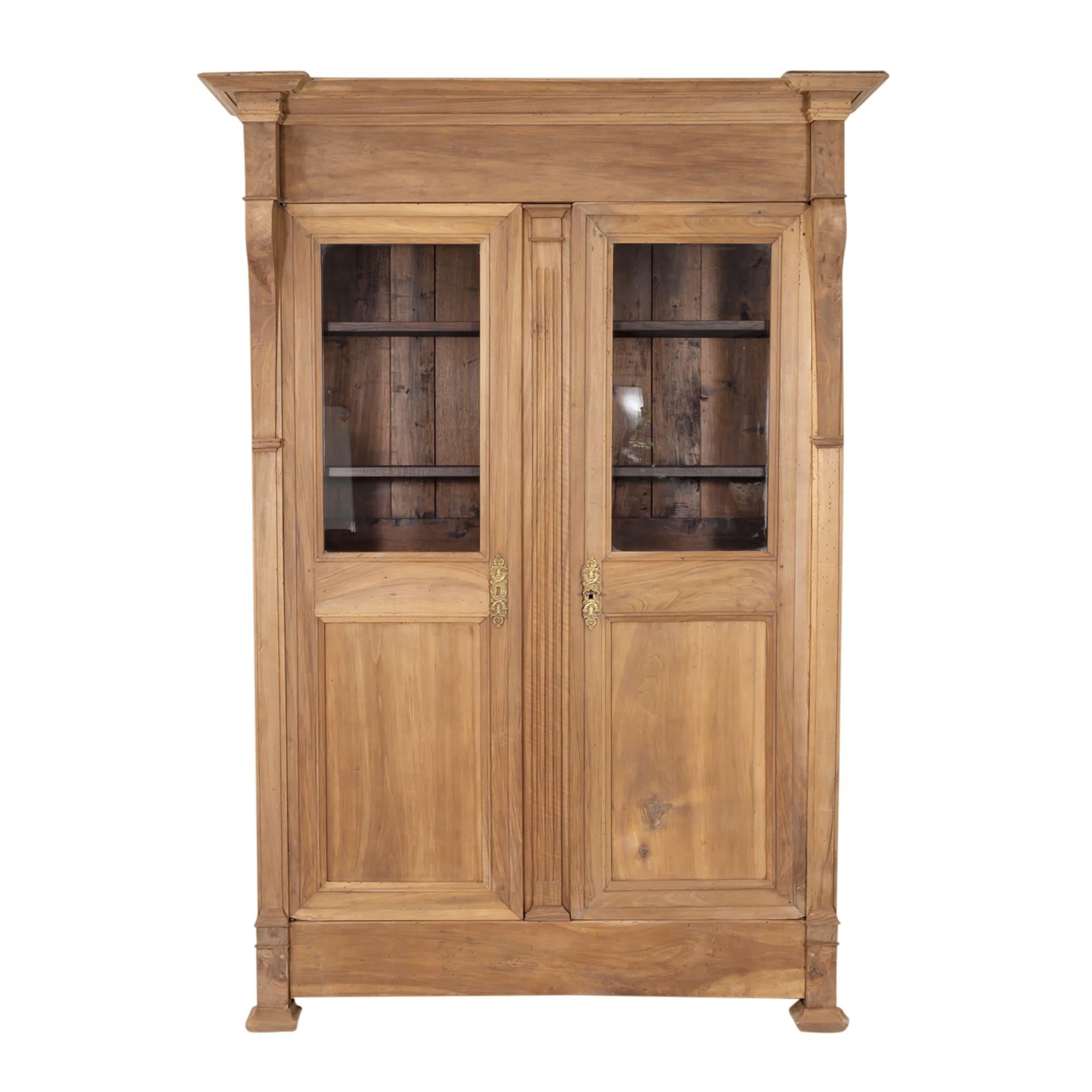 19th century French restauration period bibliotheque handcrafted of walnut in the South of France, circa 1820s. This handsome bookcase with its warm bleached patina features a stepped cornice over paneled doors with the original glass flanked by
