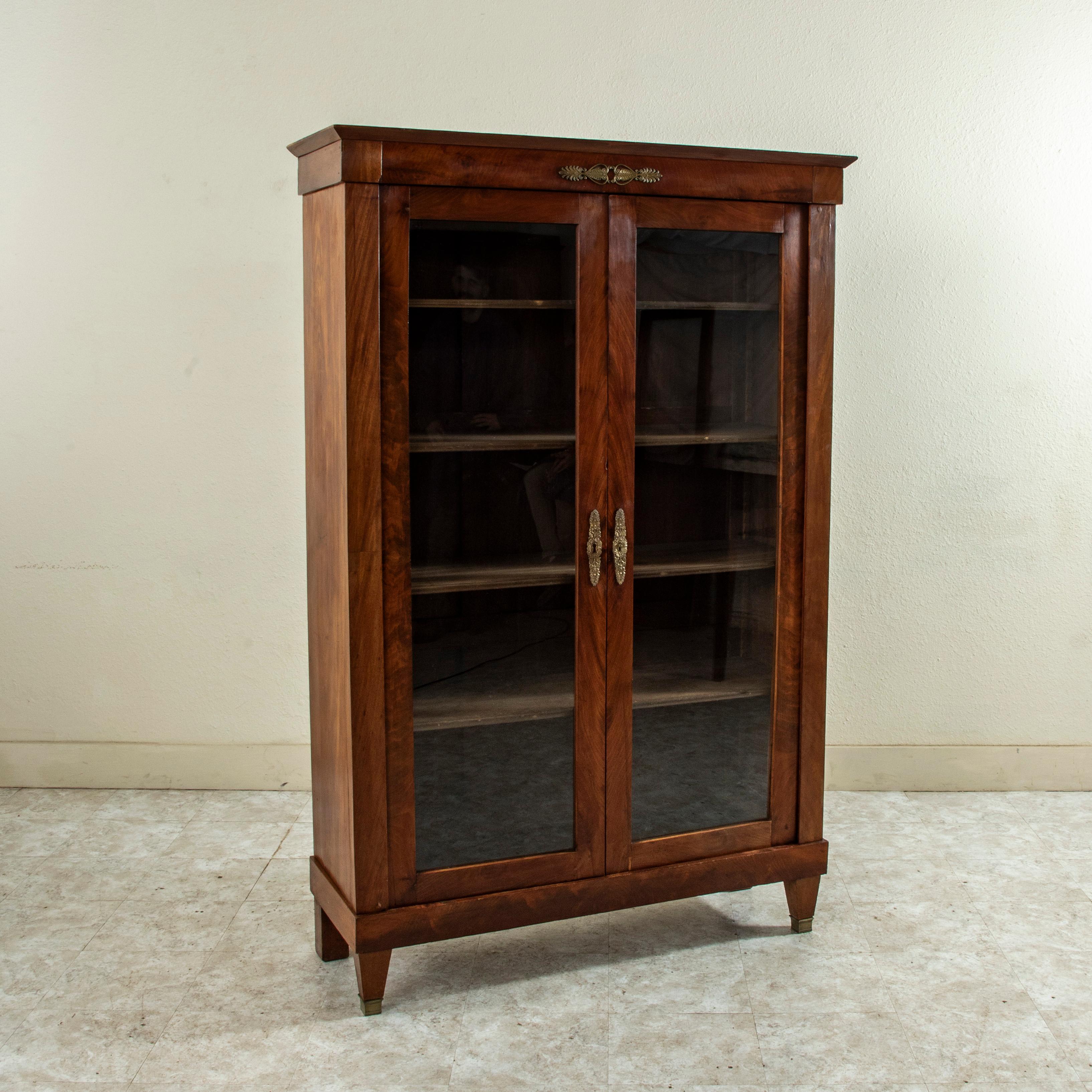 This mid nineteenth century French Restauration style bookcase or vitrine is constructed of solid mahogany and features a bronze plaque at the crown detailed with symmetrical leaves, florets, and palmettes. Its two glass doors open using a single