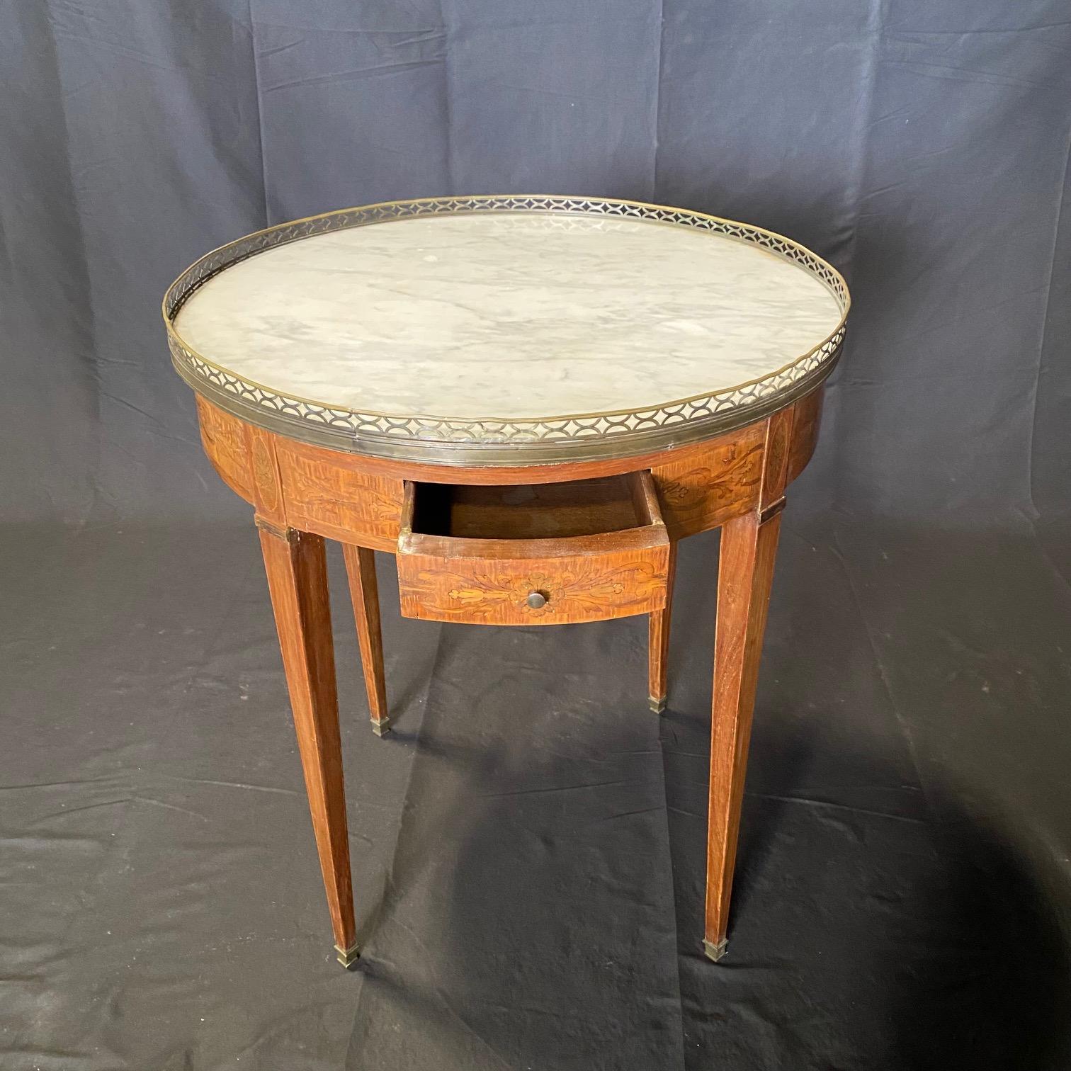 Table bouillotte ronde d'époque Louis XVI à plateau de marbre Carrera avec marqueterie de rubans élaborée. Deux tiroirs et deux tirettes ou glissières extractibles. Le plateau en marbre blanc de Carrera est entouré d'une galerie en bronze percée.