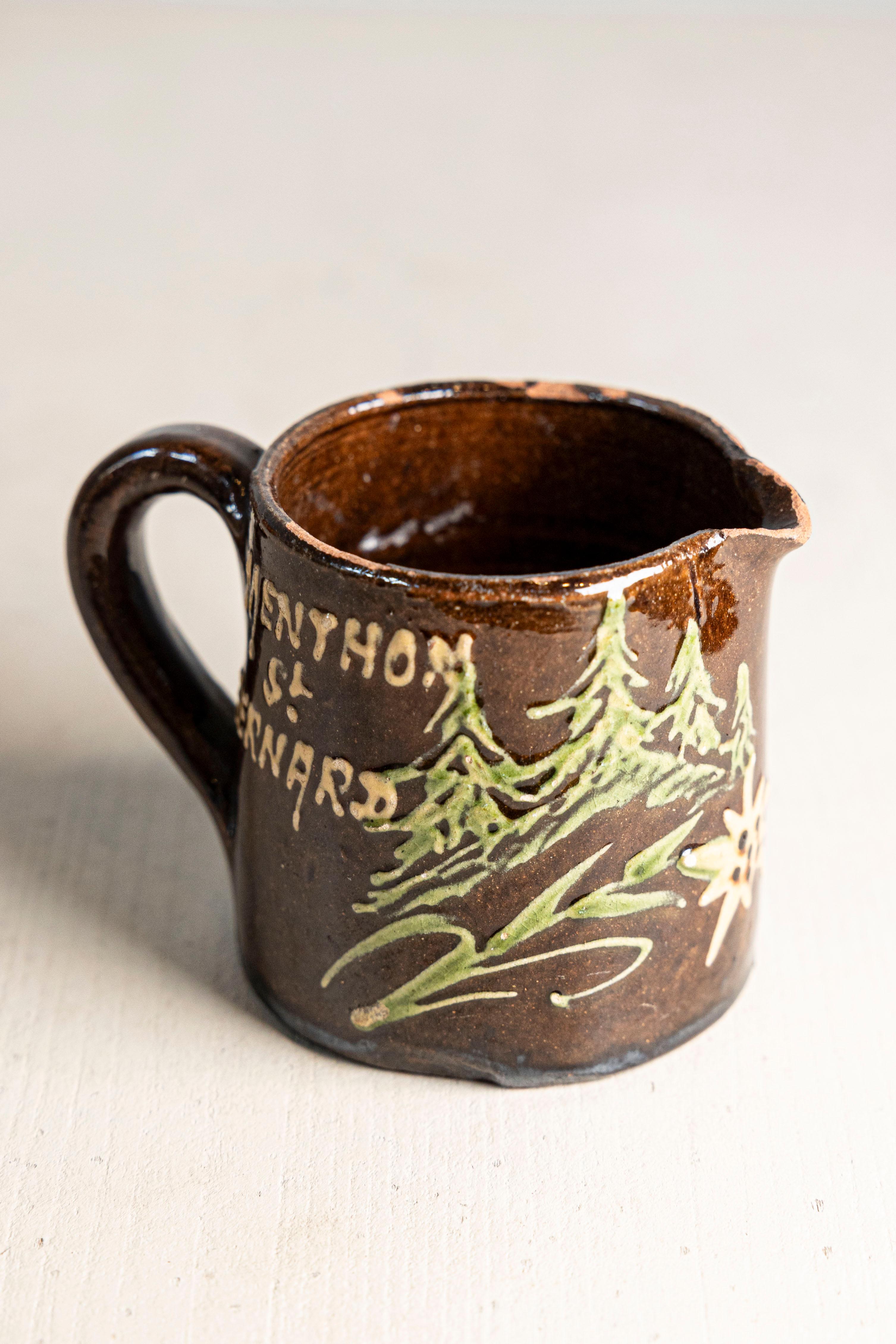 A small, rustic French souvenir pitcher from the 19th century, originating from Menthon-Saint-Bernard, a picturesque village nestled in the Haute-Savoie region near Lake Annecy. Handcrafted in terracotta and finished in a deep brown glaze, the