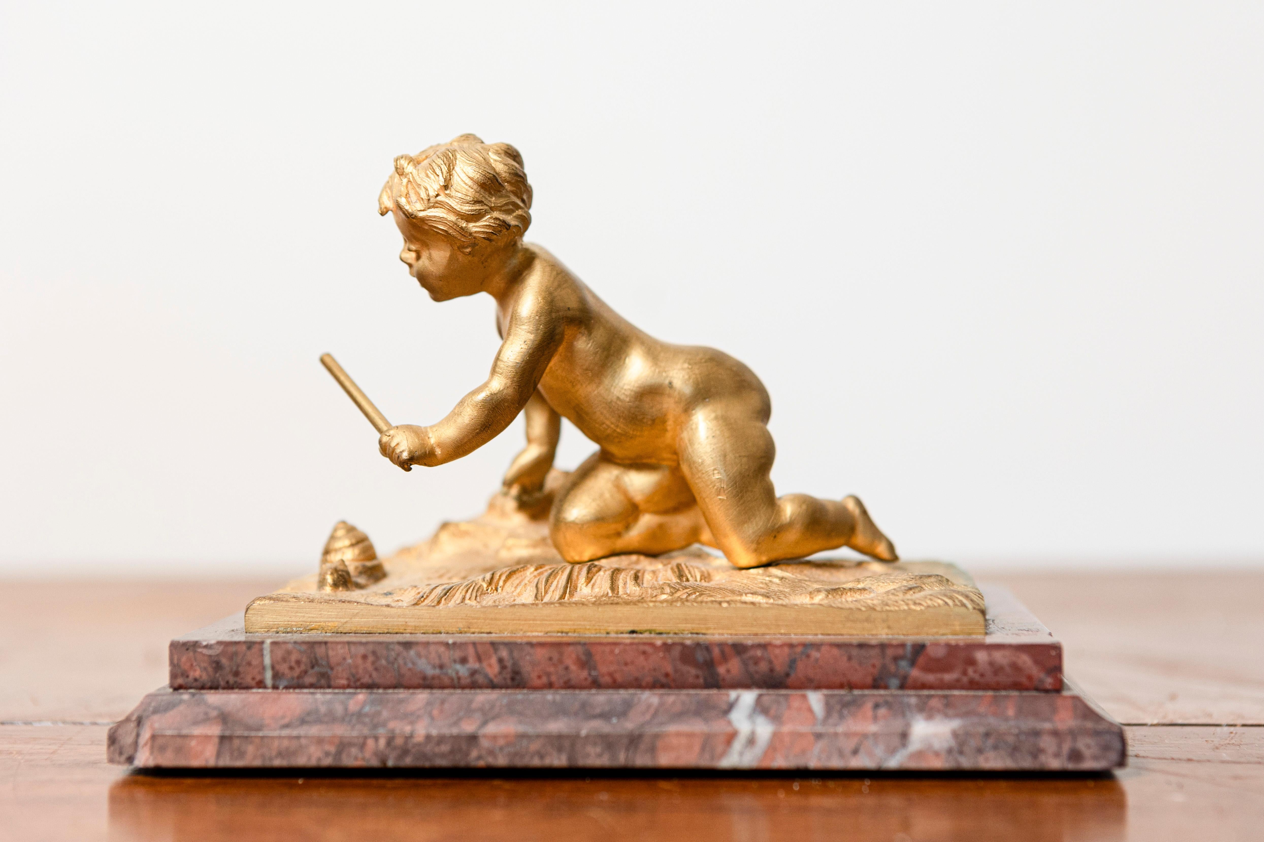 Questa scultura francese del XIX secolo raffigura un bambino in posa giocosa, inginocchiato e impegnato con un bastone, apparentemente colto in un momento di curiosità. La scultura è splendidamente realizzata in bronzo dorato e cattura l'innocenza e