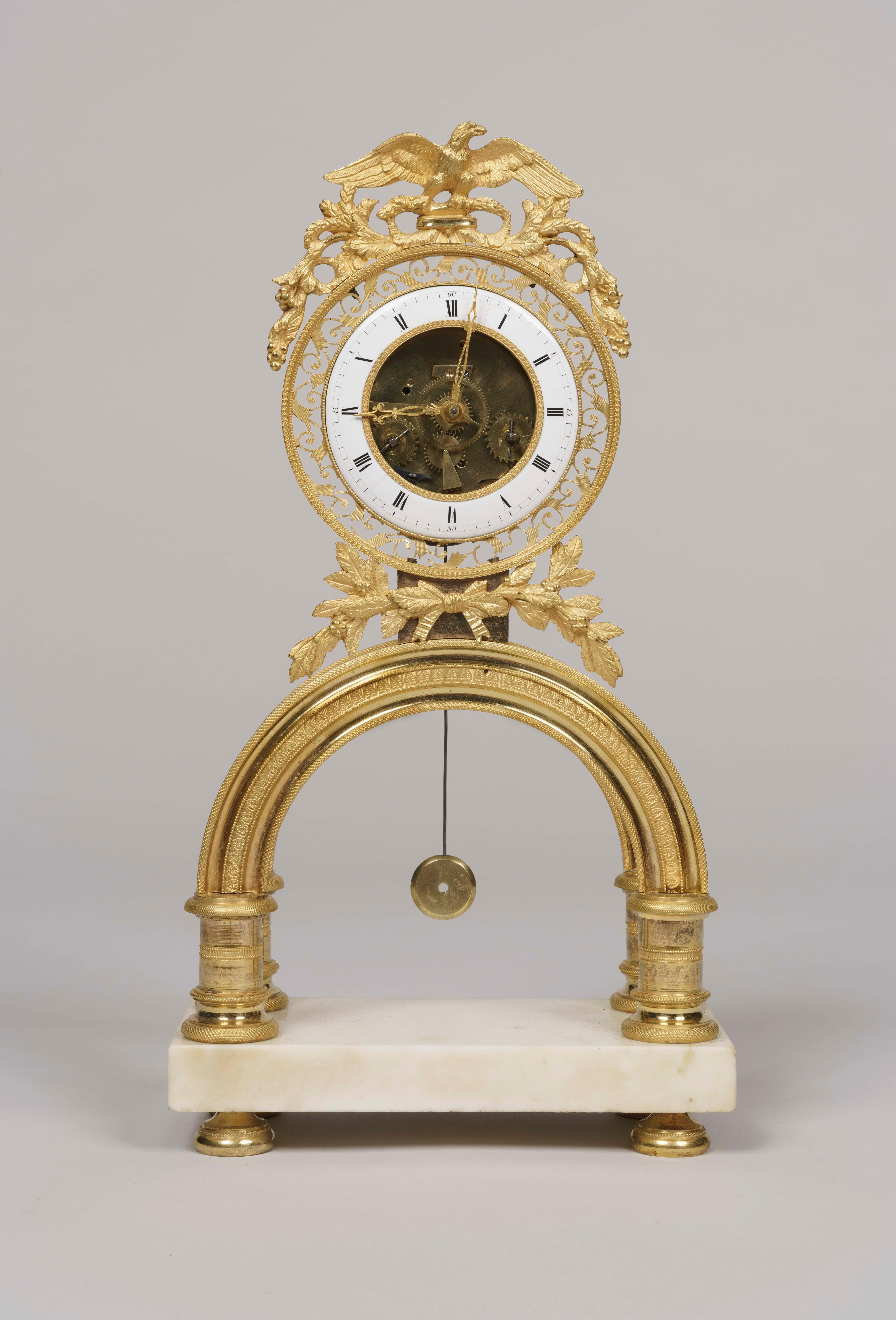 Eine symbolische Skelettuhr aus der Zeit des französischen Directoire.

Ein rechteckiger Sockel aus Carrara-Marmor mit Bronzefüßen mit Rändeldekoration trägt einen Bogen aus Ormolu, über dem sich die runde, mit Girlanden geschmückte Uhr befindet.