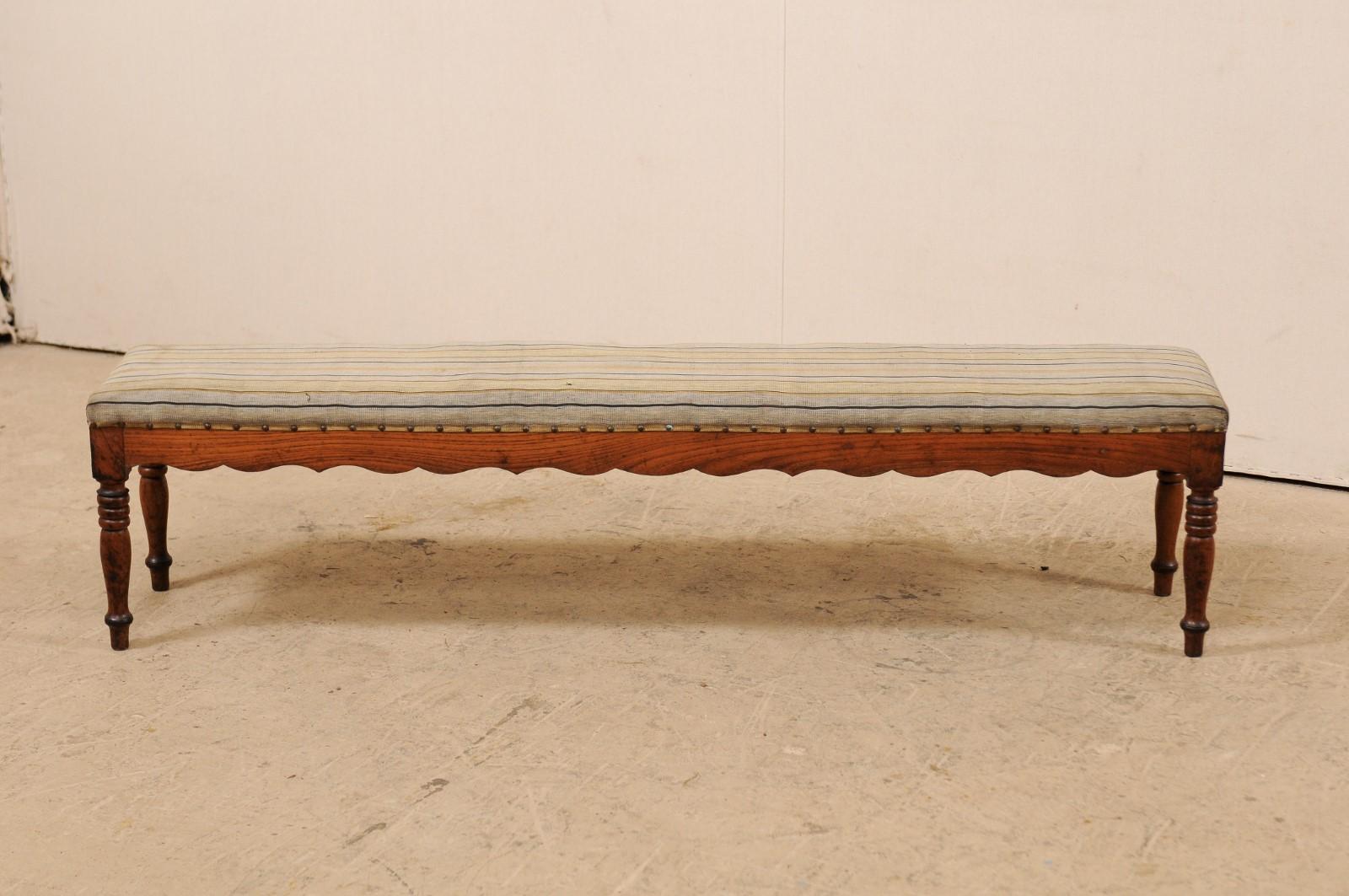 A slender French upholstered seat and carved wood bench from the early 19th century. This antique bench from France features a thinly profiled and upholstered seat, with a wood skirt with wavy carved designs about all bottom sides, and is raised