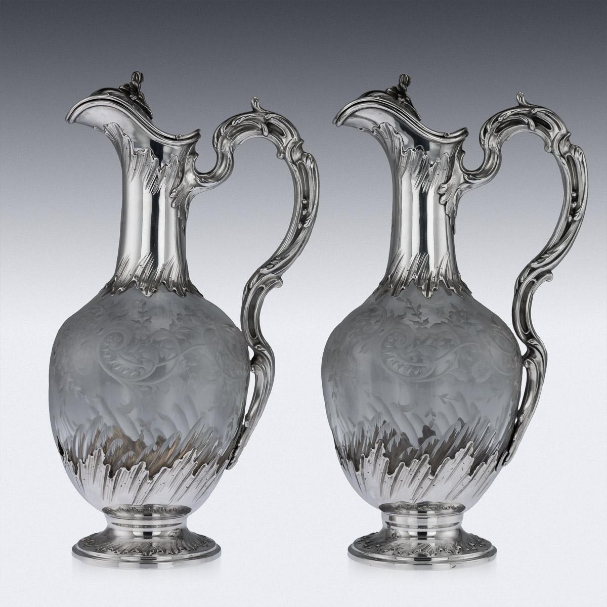 Antique late 19th century French solid silver and glass pair of claret jugs, each body is baluster form, beautifully cut and etched, the silver foot, neck and handle are chased with leaves and scrolls, the hinged lid is surmounted by a cast floral