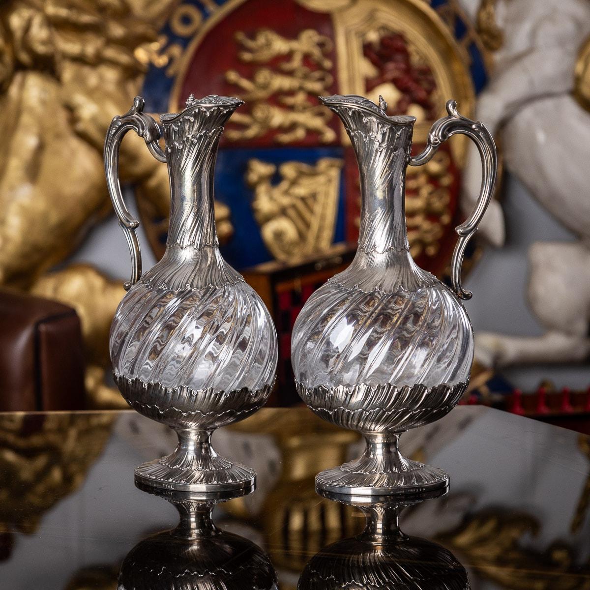 Antique late 19th century French solid silver and clear glass pair of claret jugs, each body is of baluster form, beautifully swirled glass, the applied silver foot, neck and scroll handle are chased with leaves and scrolls, the hinged lid is