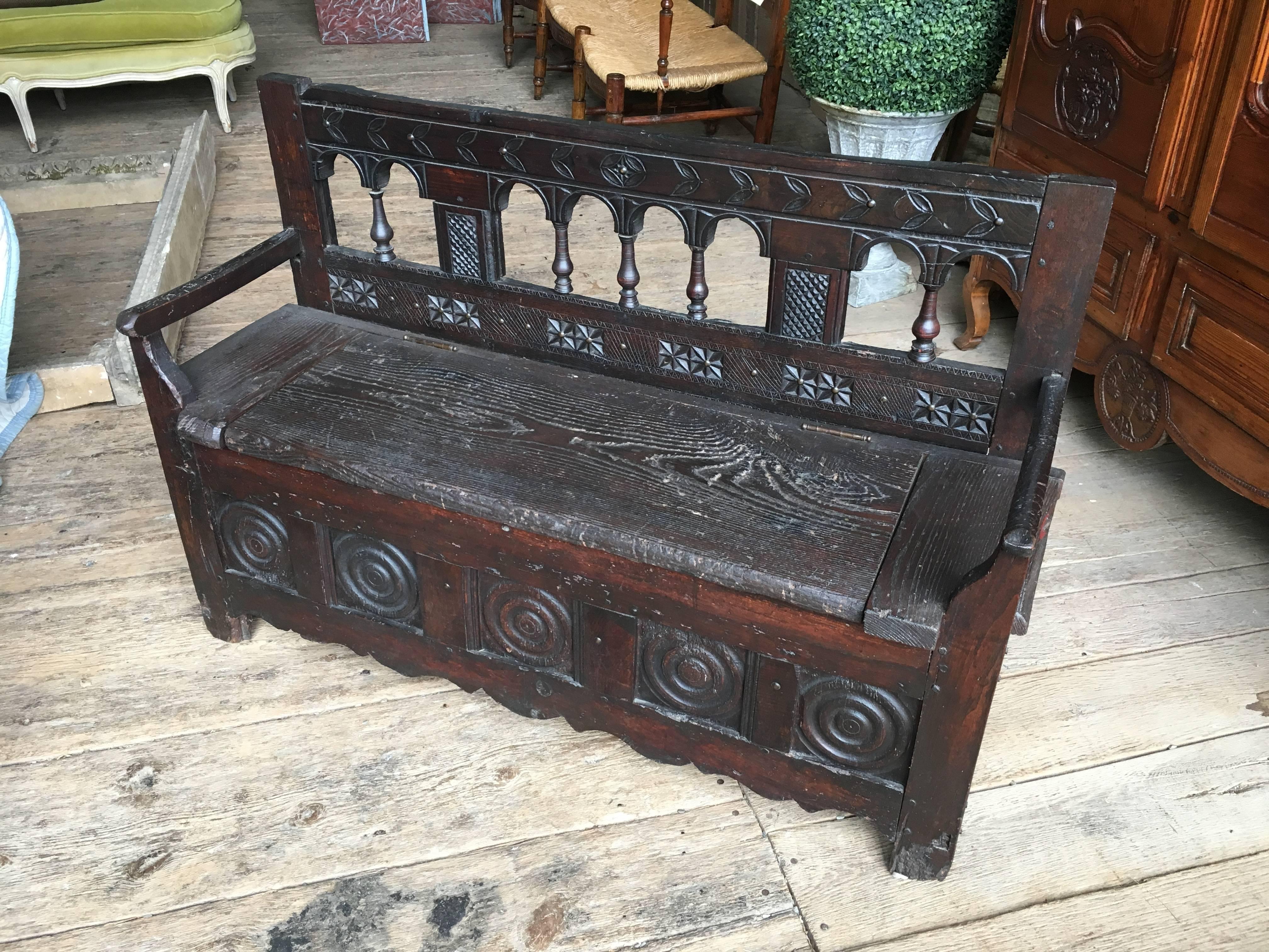 A rustic 19th century storage bench in dark oak from Brittany France. The back is decorated with a carved top rail with arches and turned balusters. The seat lifts to a storage compartment with arms on both sides. The base is scalloped with circular