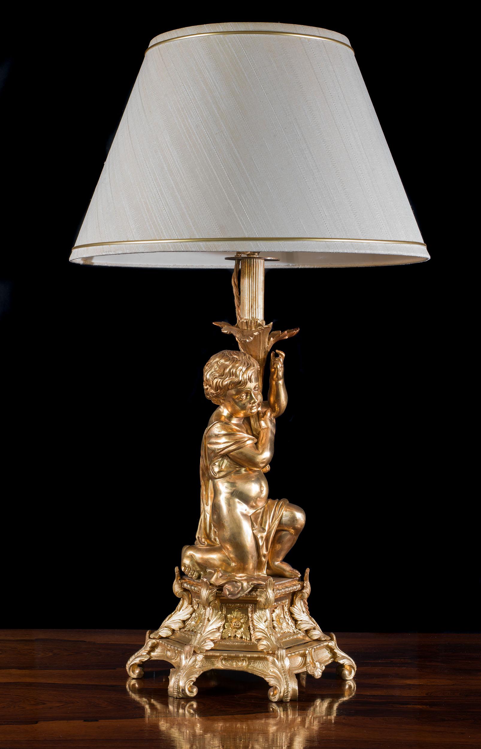 A large and ornate gilt bronze lamp, modeled as a putto holding the lamp fitting aloft whilst kneeling on a hexagonal base embellished with foliate Rococo decorations.

French, c. 1880.