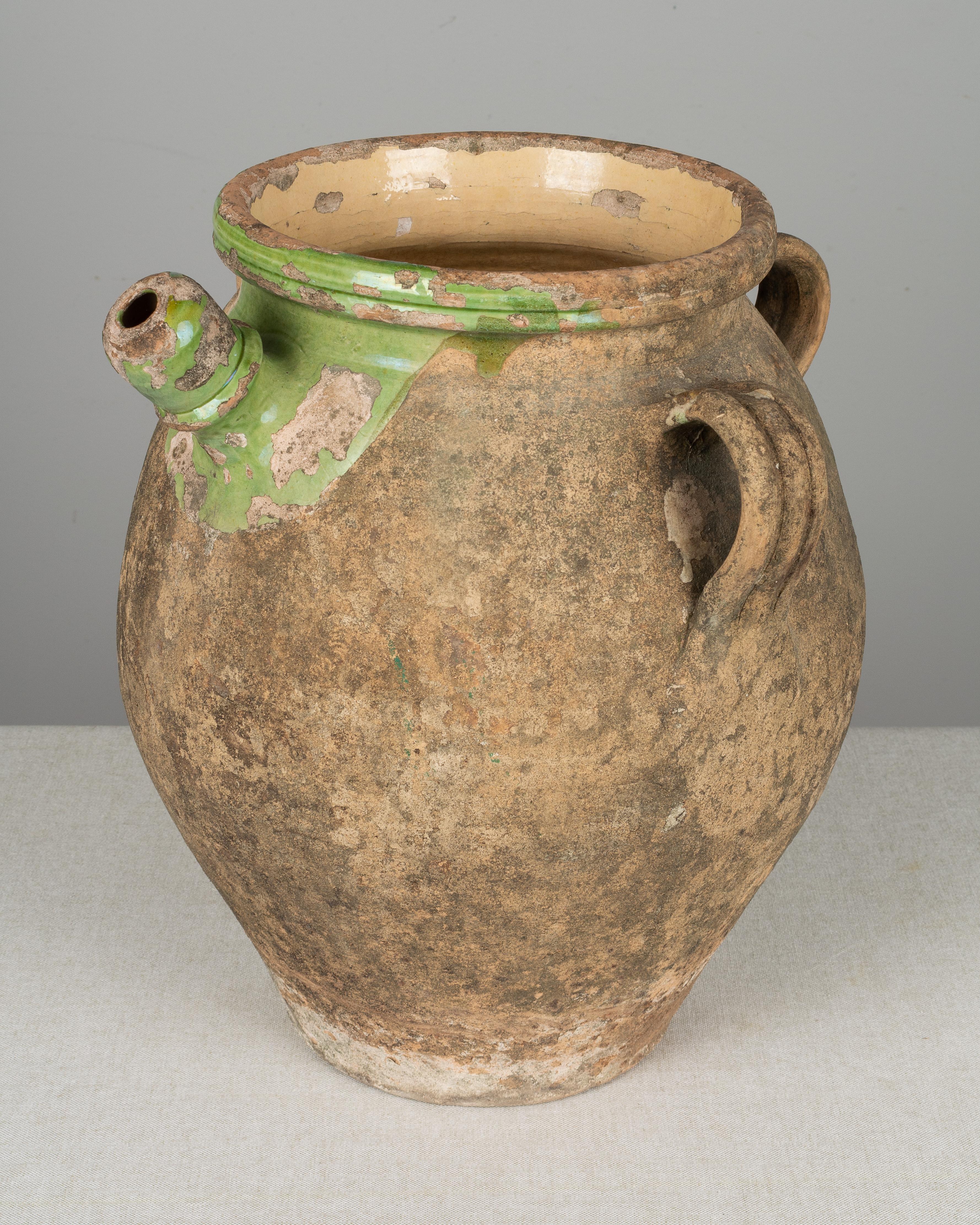 A 19th century earthenware pot from the Southwest of France with three handles and a large spout. Old weathered patina with remnants of pale jade green glaze on the exterior and pale yellow glaze on the interior. There is a hole in the bottom for