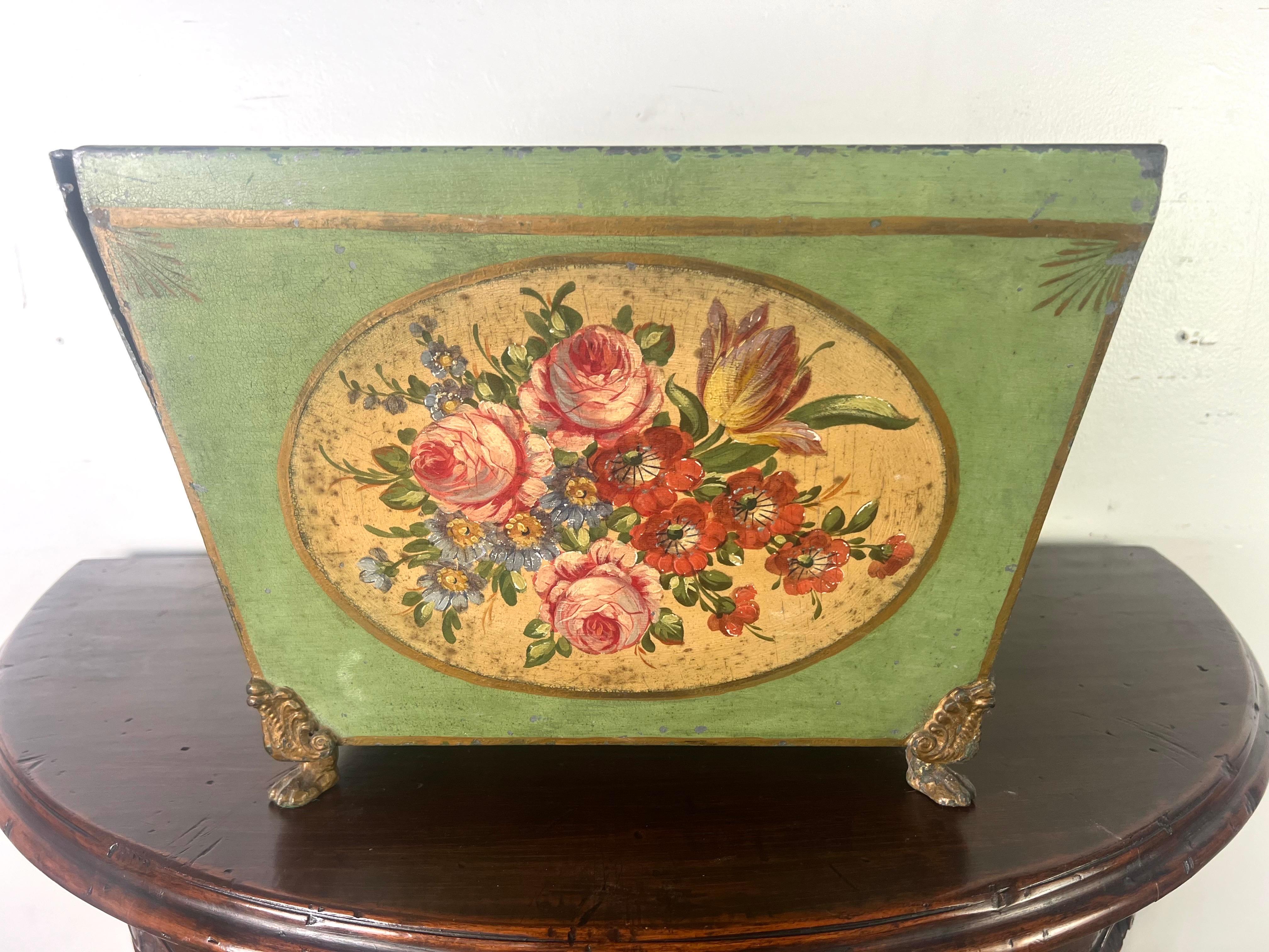 19th-century French tole container.  It features a trapezoidal shape and stands on four ornate brass feet with acanthus leaf motifs.  The main body is adorned with a bouquet of flowers painted in a palette of gold, reds, yellows, pinks, glues and